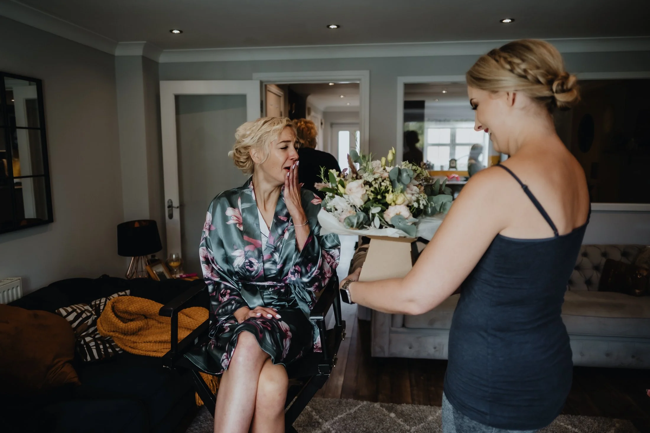 A woman in a floral robe is sitting on a chair, emotional and crying, while another woman in a black dress offers her a bouquet of flowers in a cozy living room.