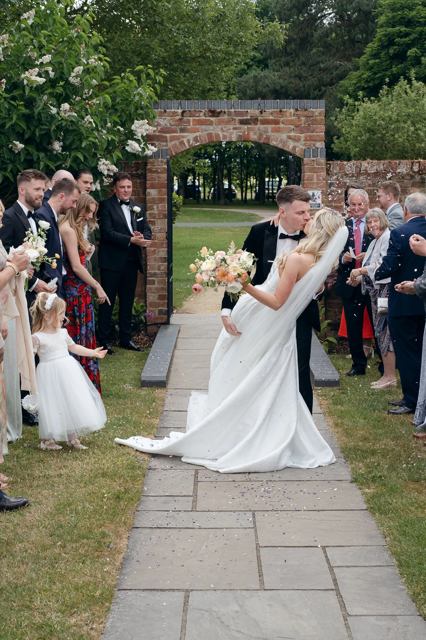 A bride and groom share a kiss outside at their wedding ceremony, surrounded by wedding guests, flower arrangements, and greenery.