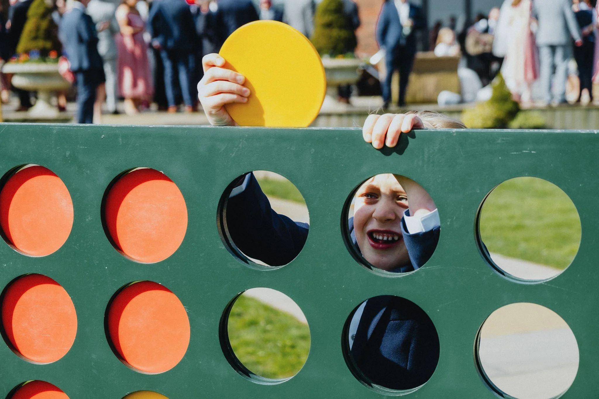 A child playing connect four outdoors, holding a yellow disc, with other children and adults in the background.