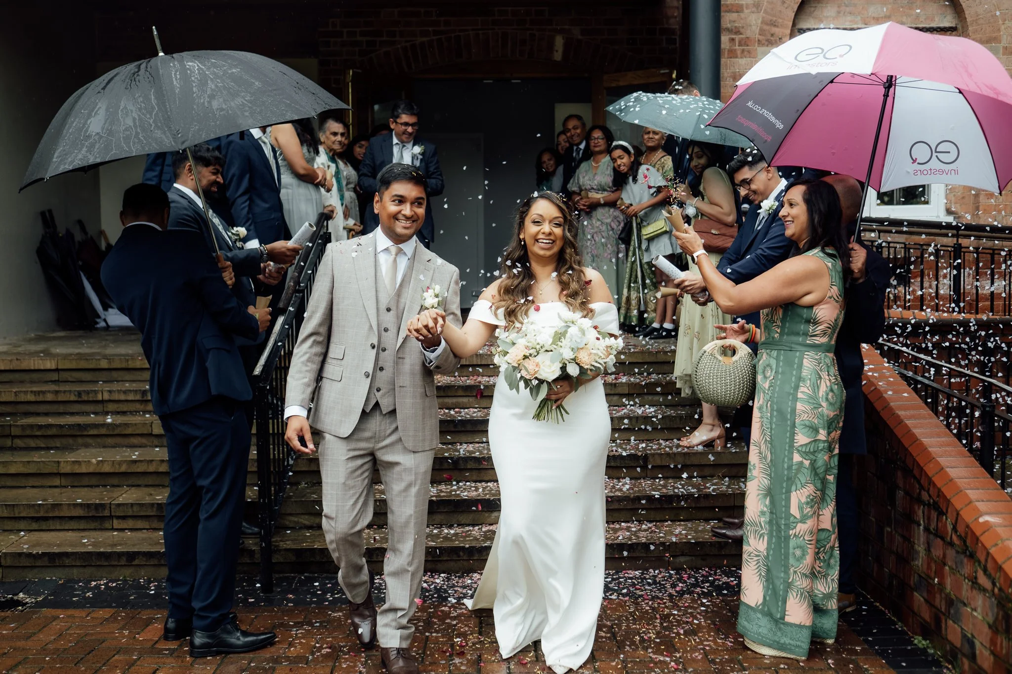 A newlywed couple walking down a brick staircase, holding hands, surrounded by friends and family in formal attire, celebrating and throwing confetti on a rainy day with umbrellas.