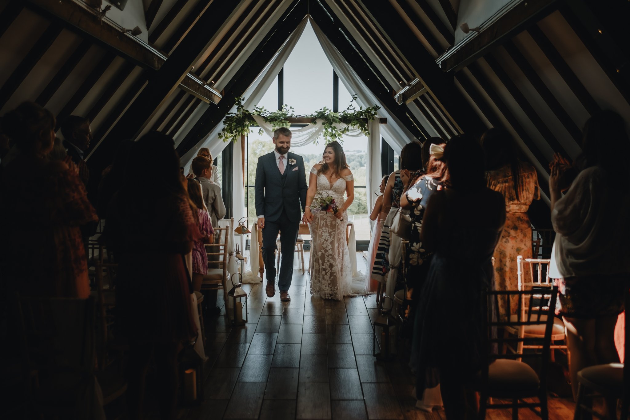 A bride and groom walking down the aisle during their wedding ceremony inside a wooden A-frame chapel with large windows, decorated with greenery and white drapery, with guests standing on either side watching and clapping.