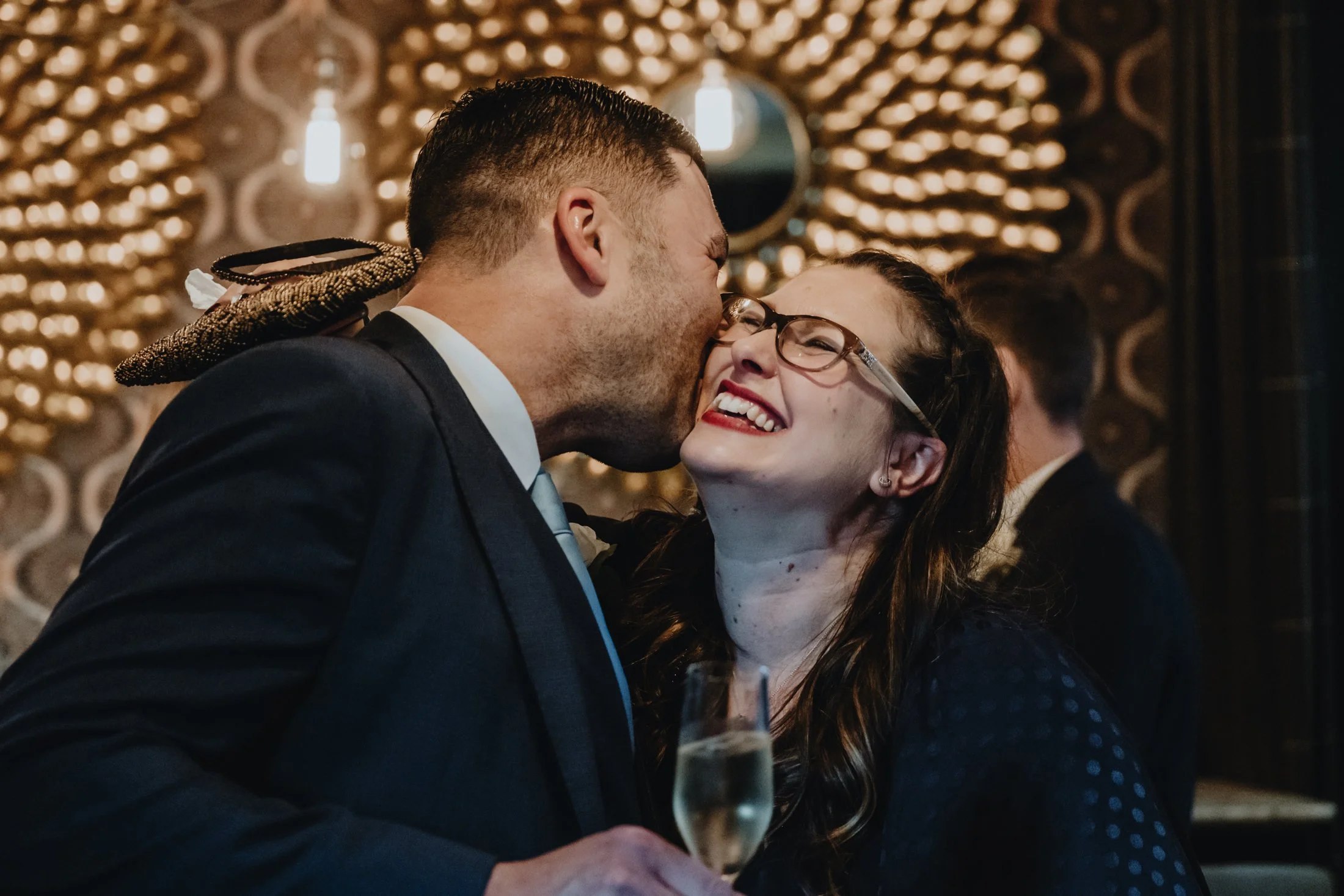 A man and woman sharing a joyful kiss at a celebration or party, with the woman wearing glasses and a dark dress, and the man in a suit. The woman is smiling broadly and holding a glass of champagne. The background is decorated with warm lighting and