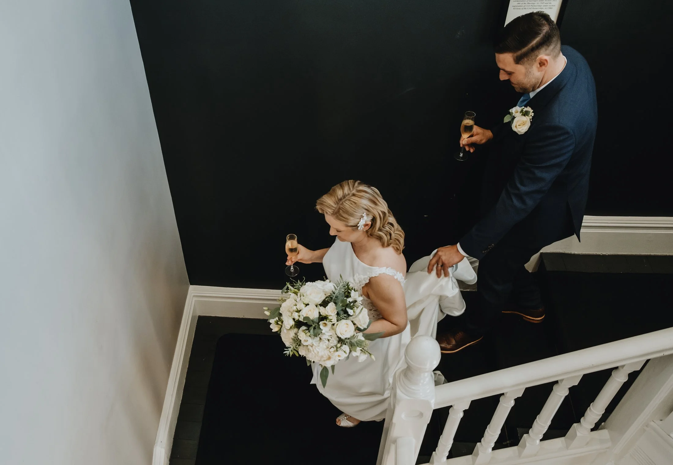 A bride walking down a staircase holding a bouquet of white flowers, a groom in a dark suit with a boutonnière holding a champagne glass, captured from an overhead angle.