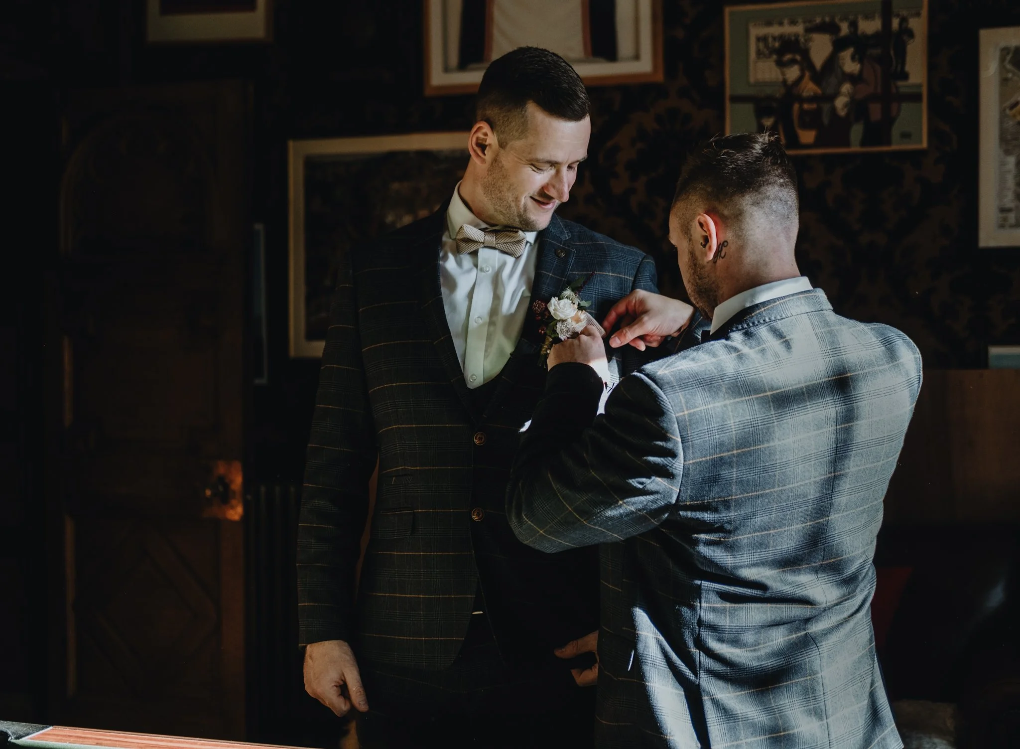 Two men dressed in suits, one is pinning a boutonniere on the other's lapel in a warmly lit room decorated with framed artwork.