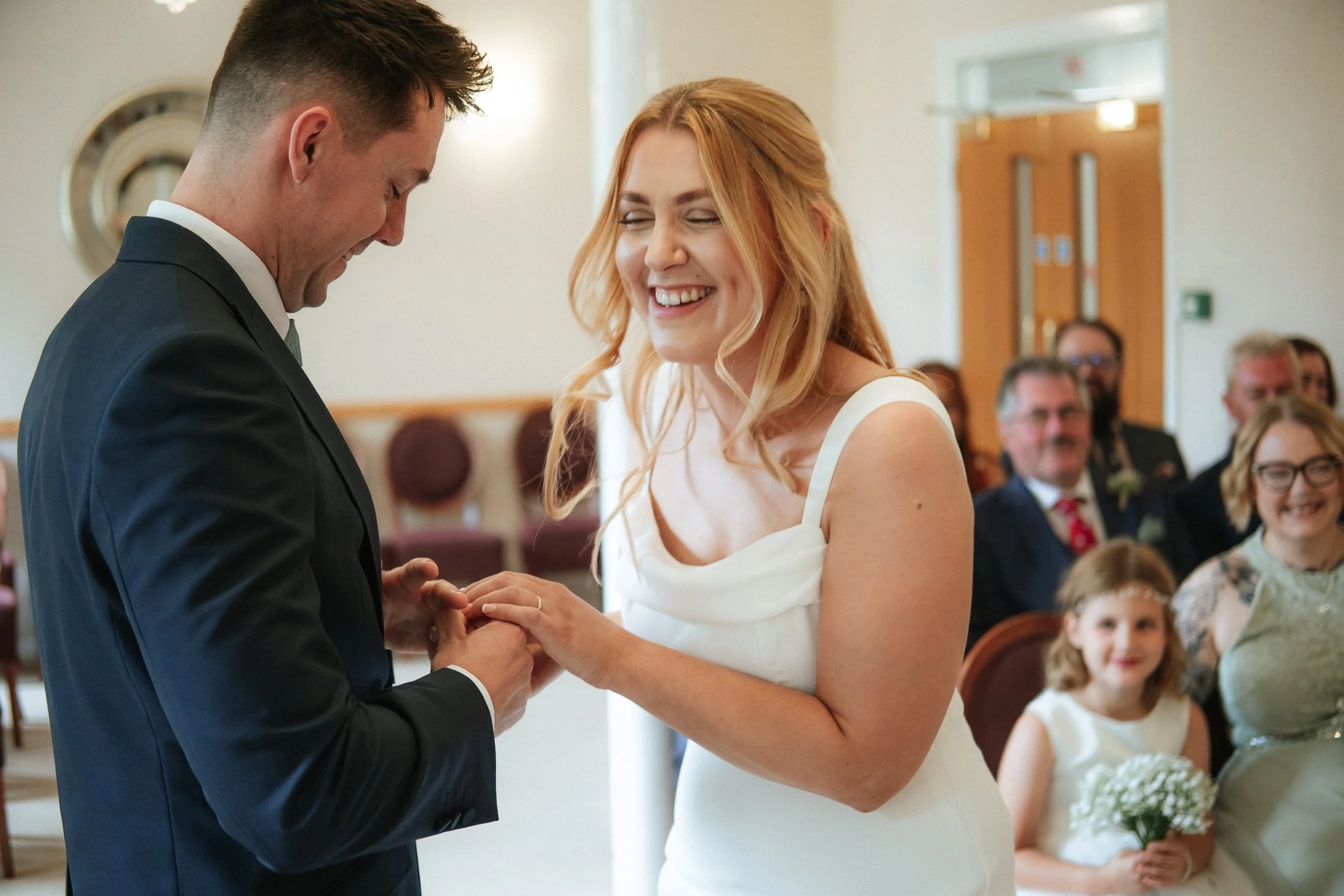 A couple during their wedding ceremony, with the groom placing a ring on the bride's finger, both smiling happily with guests in the background.
