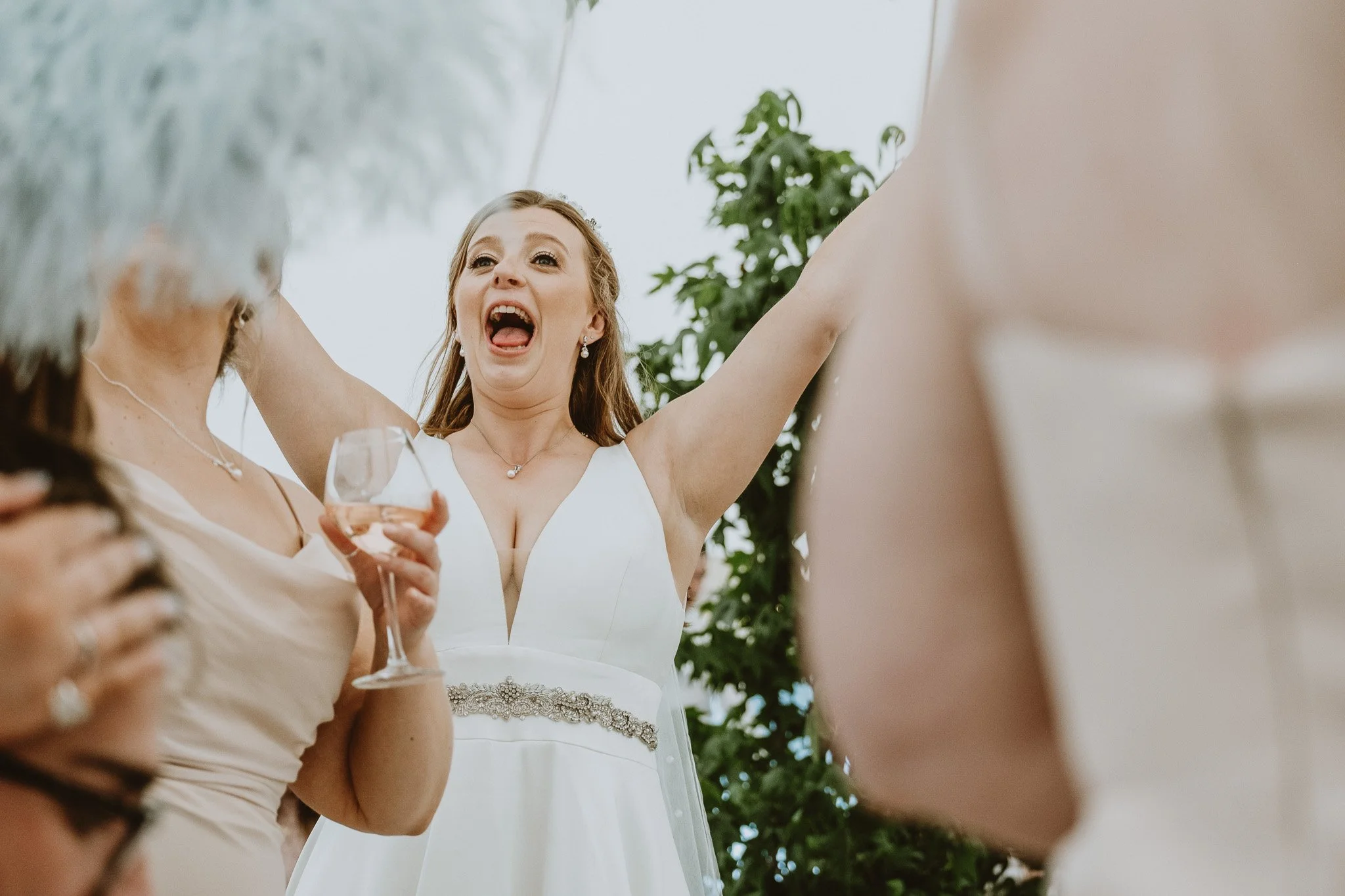 A woman in a white dress celebrating, with her arms raised and a joyful expression at a wedding reception.