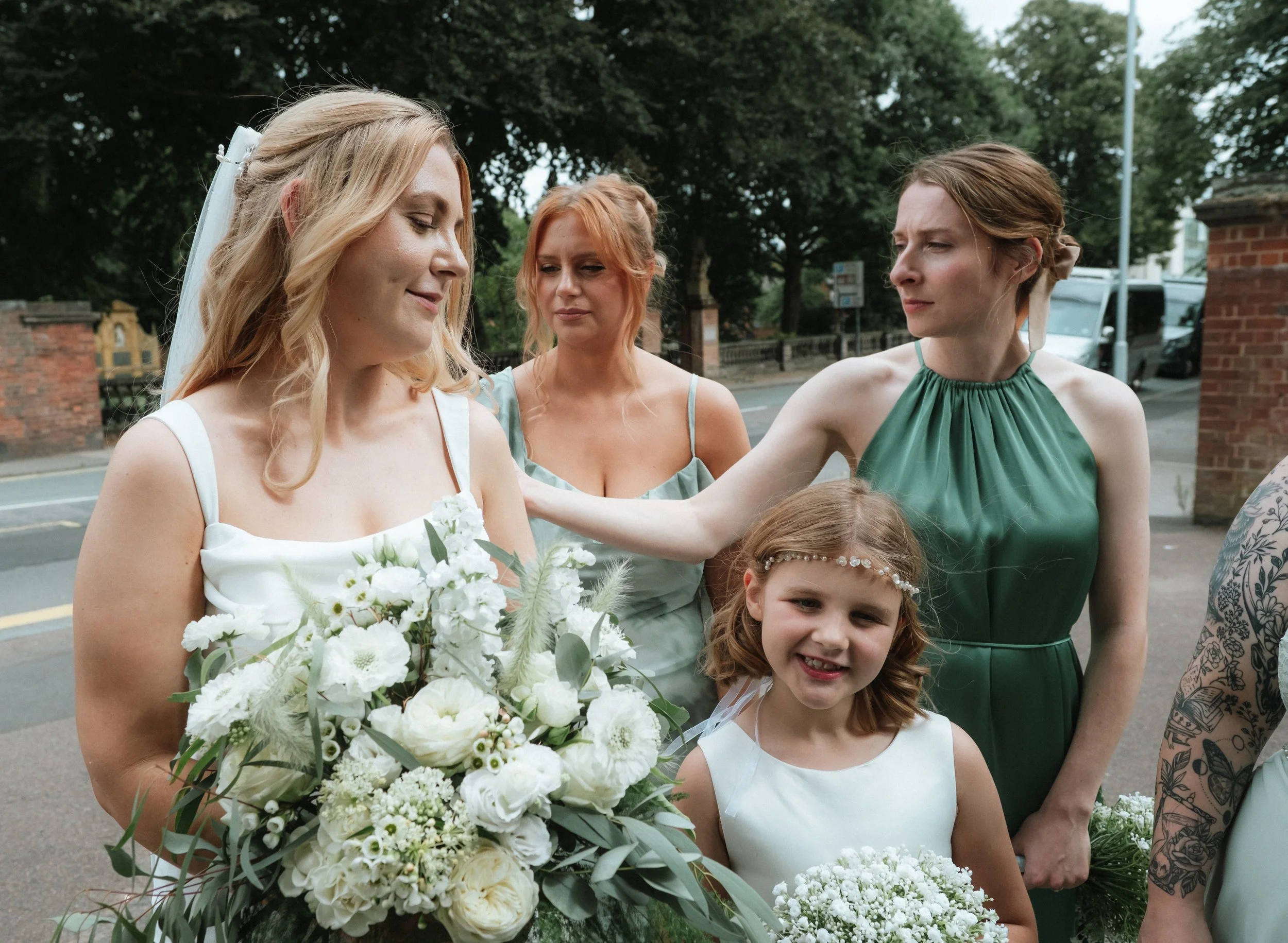 A bride holding a large bouquet of white flowers standing outdoors with four women and a young girl, with trees and a street in the background.