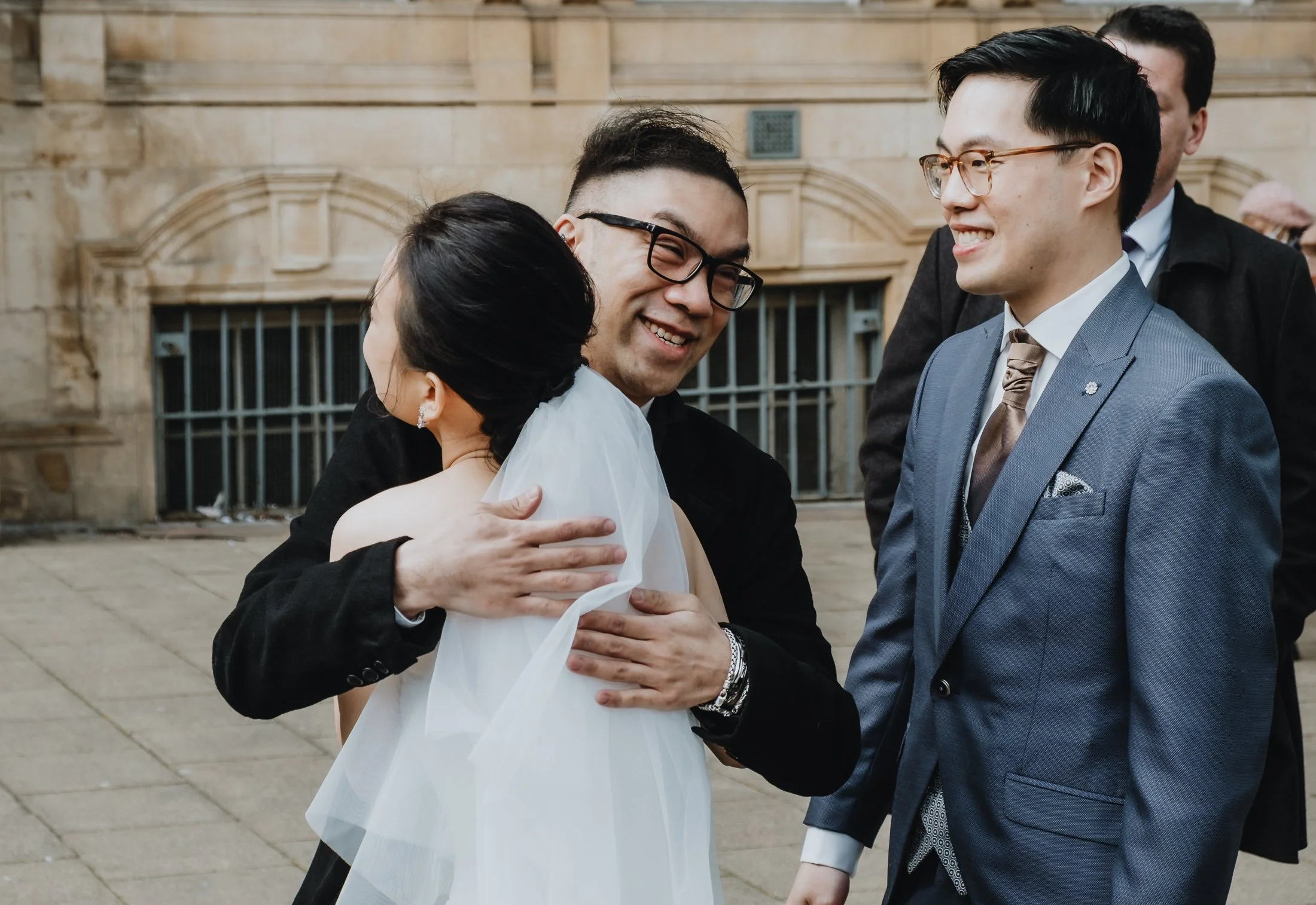 People embracing and smiling at a wedding outside a historic building.