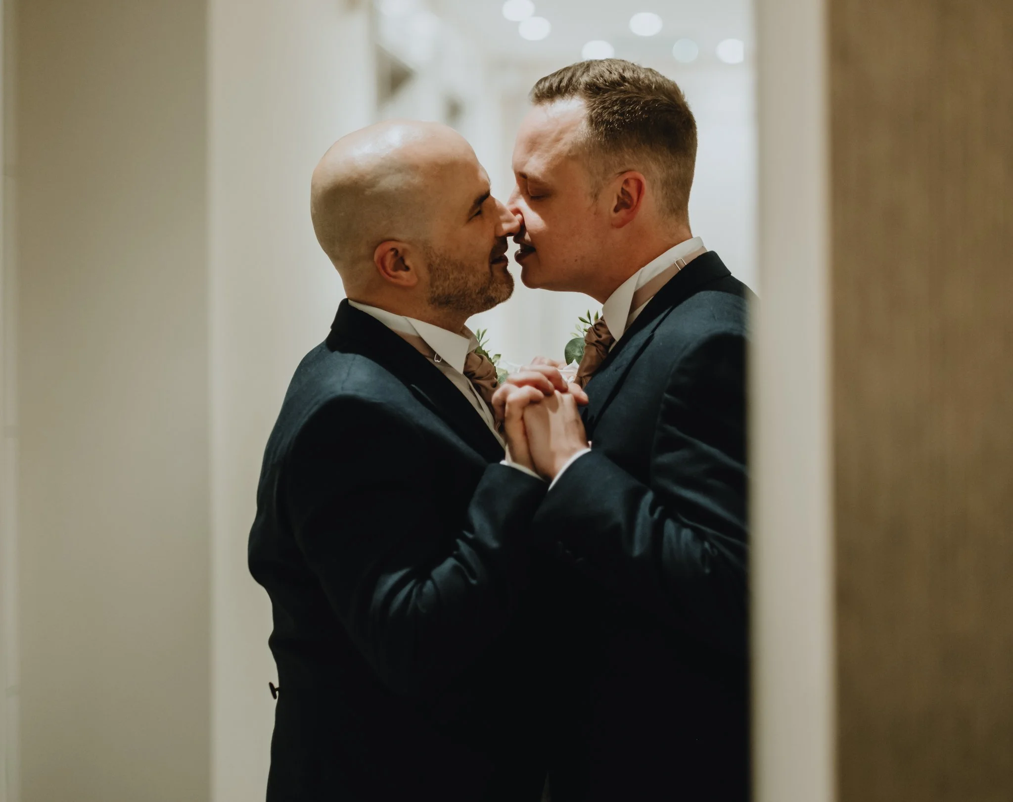 Two men in tuxedos holding hands and touching foreheads in an intimate moment.