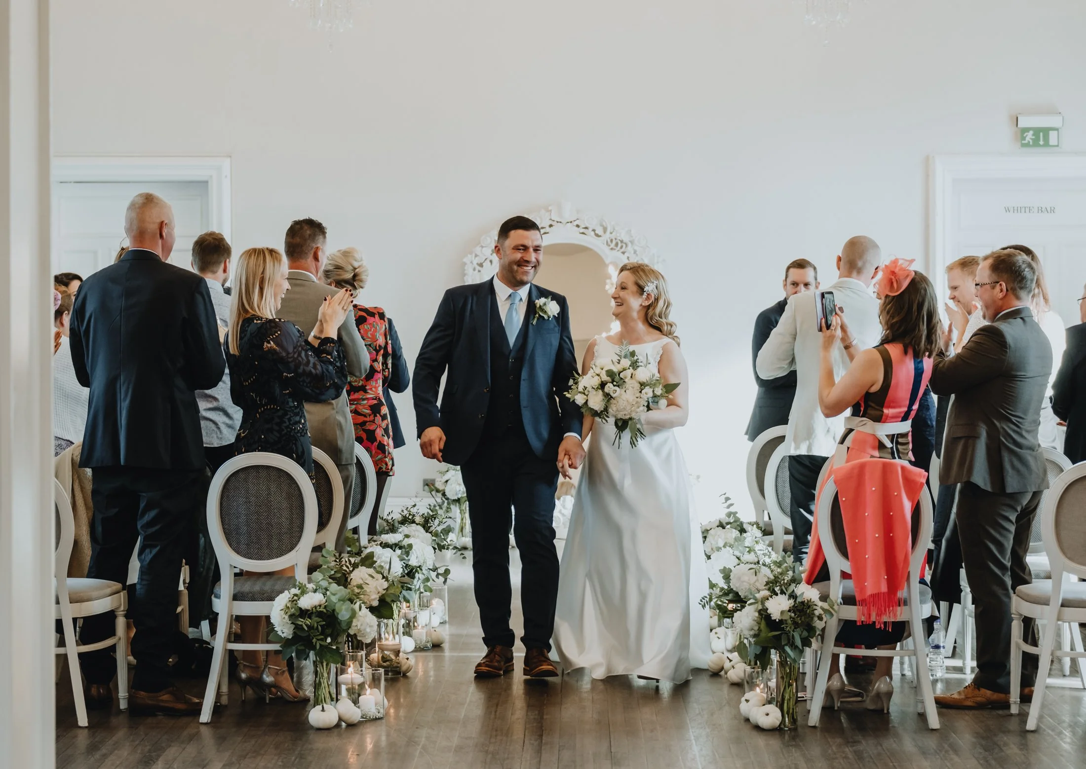 Couple walking down the aisle during their wedding ceremony, surrounded by guests, floral decorations, and candles, in a bright indoor setting.