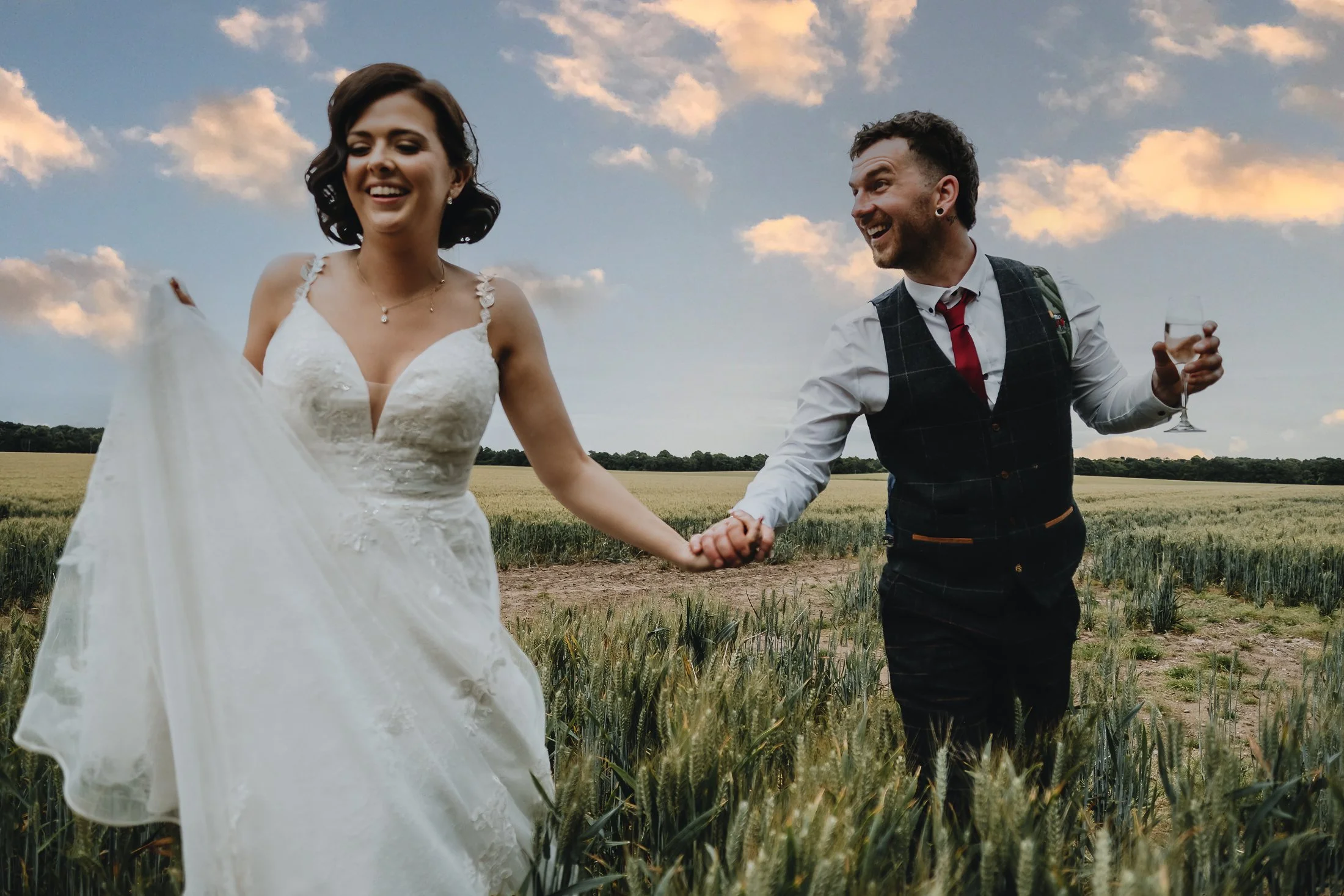 A newlywed couple holding hands and running through a field of tall grass at sunset, with the woman in a white wedding dress and the man in a vest, dress shirt, and red tie, both smiling and enjoying the moment.