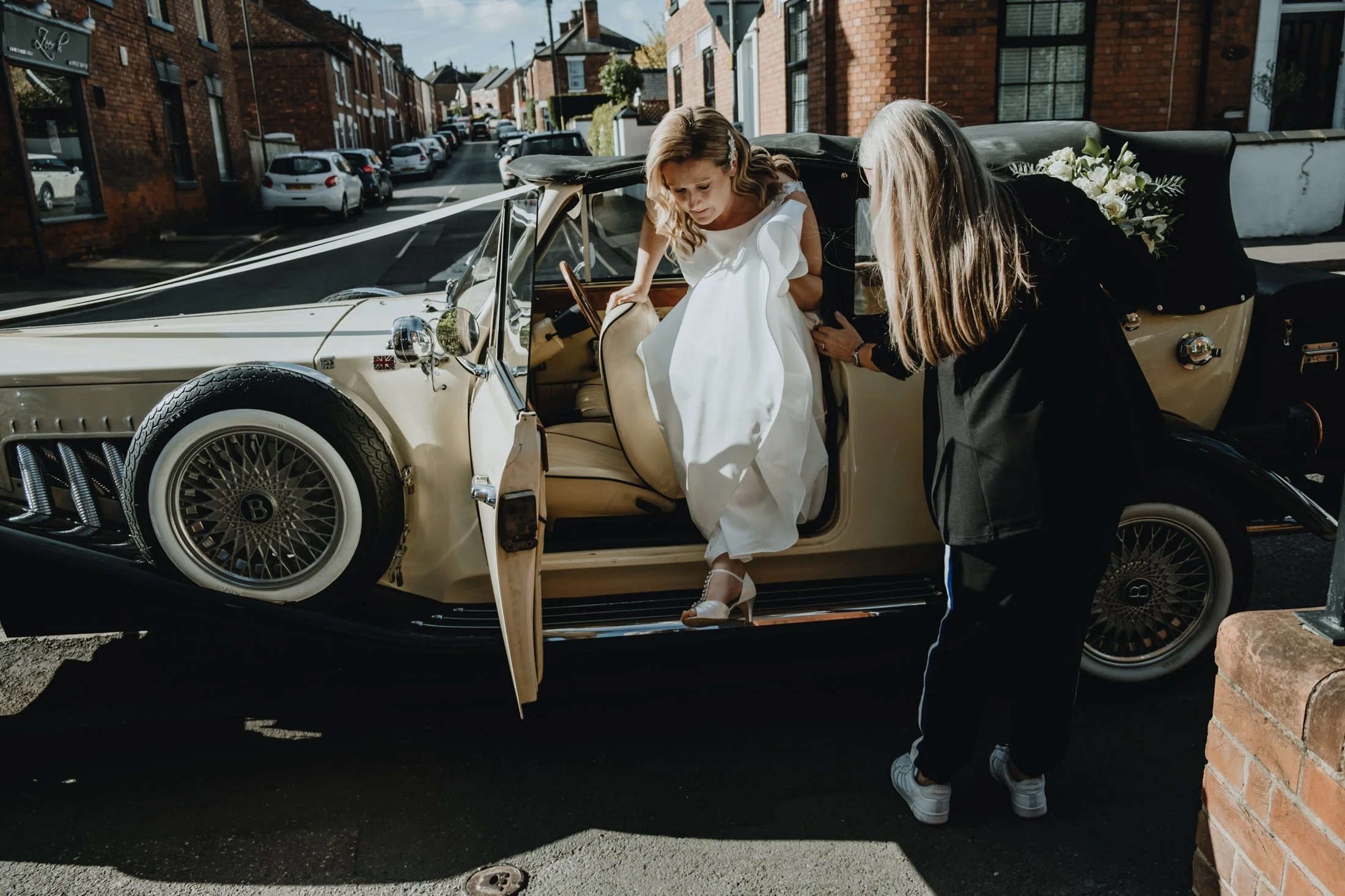 A bride in a white wedding dress stepping out of a vintage cream-colored car, assisted by a woman with long blonde hair, while on a city street with brick buildings and parked cars in the background.
