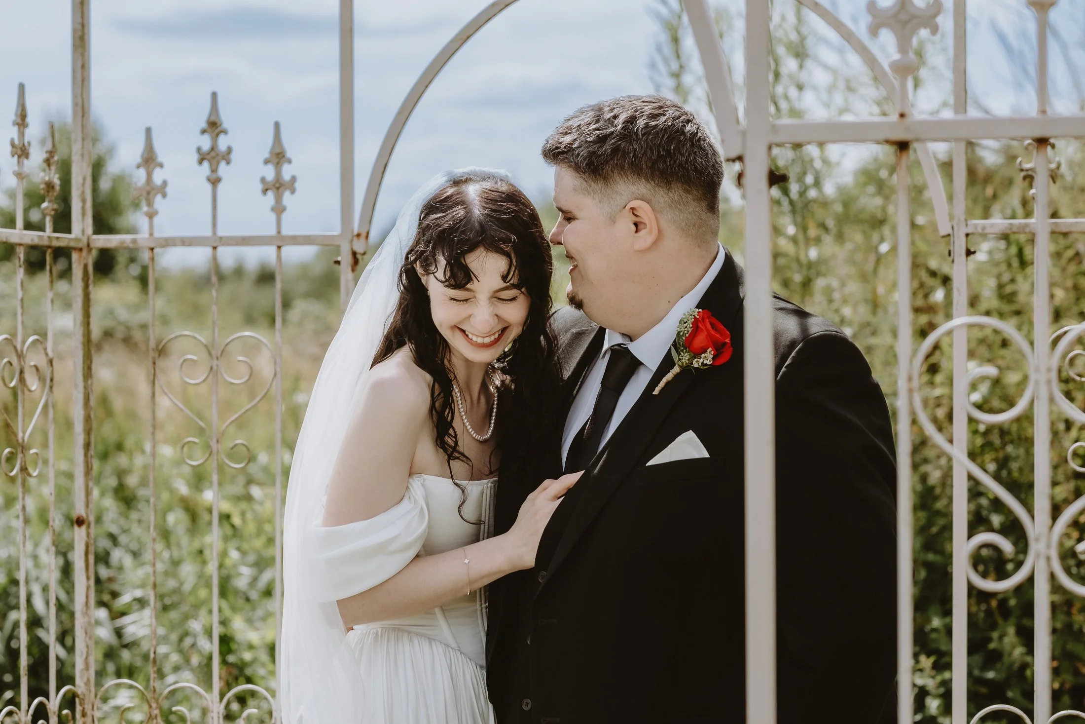 A bride and groom smiling and touching each other through a decorative iron gate outdoors on a cloudy day.