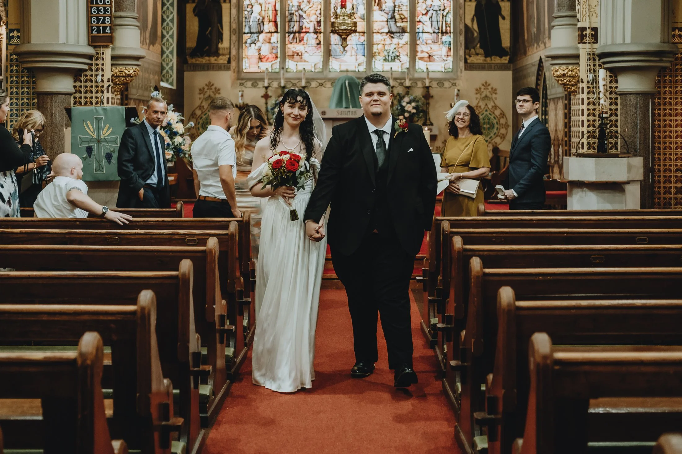 A newly married couple walking down the aisle in a church, the bride holding a bouquet of red and white flowers and wearing a white wedding dress, and the groom dressed in a black suit. Guests are standing and watching in the background.