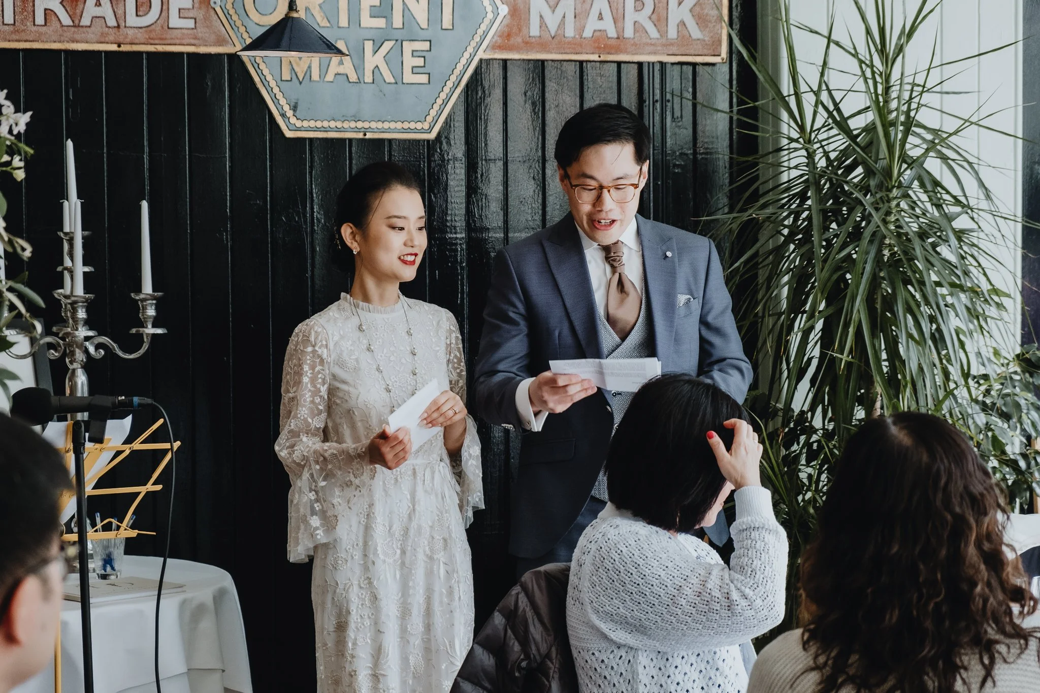 A man in a gray suit giving a speech at a wedding reception, standing next to a woman in a white lace dress who is holding a piece of paper, with seated guests listening. There are plants and a black wall in the background, and a table with a white c
