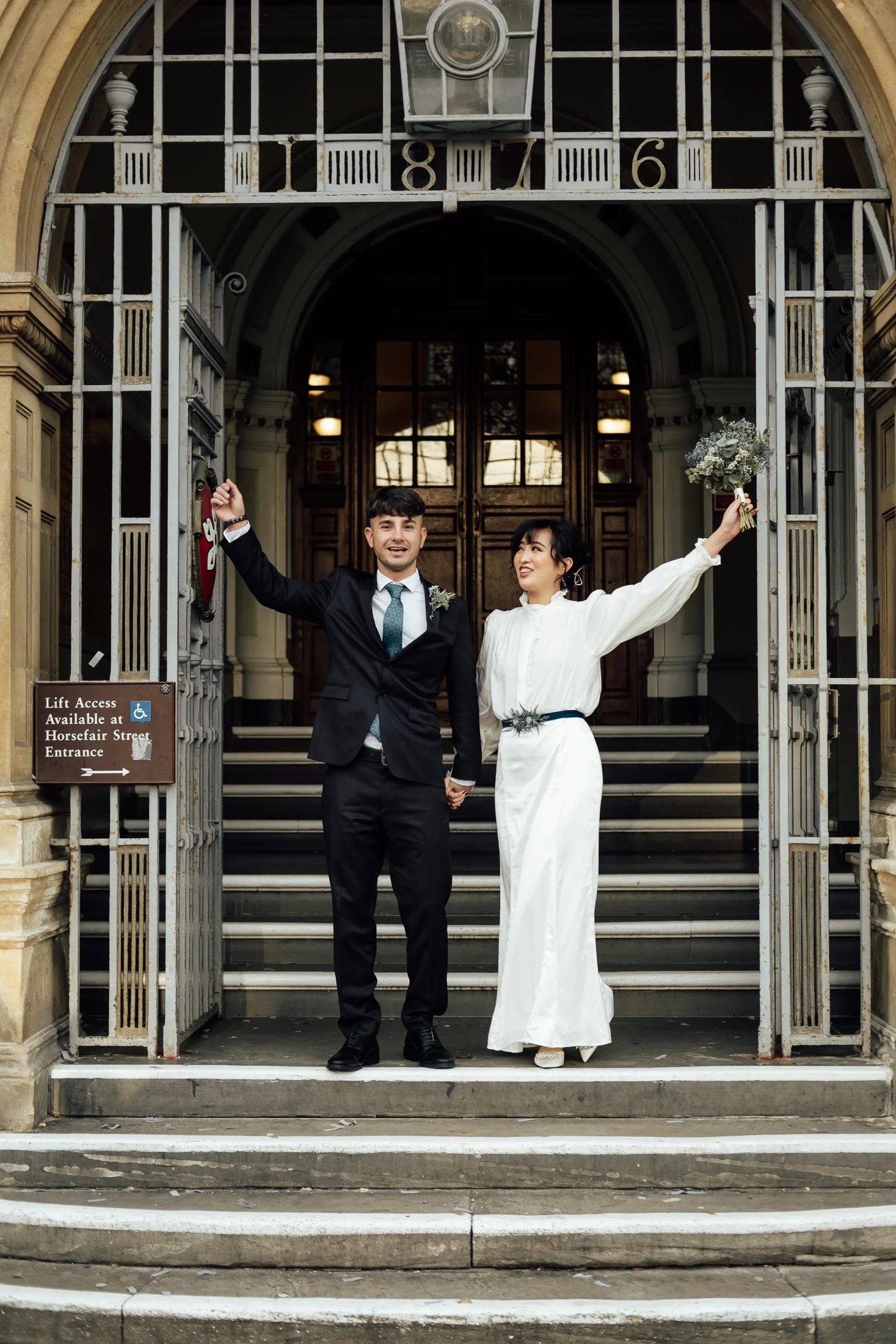A newlywed couple standing on the steps of a building with an iron gate, holding hands and celebrating. The groom is in a black suit with a blue tie, and the bride is in a white dress with her arm raised, holding a bouquet of flowers.