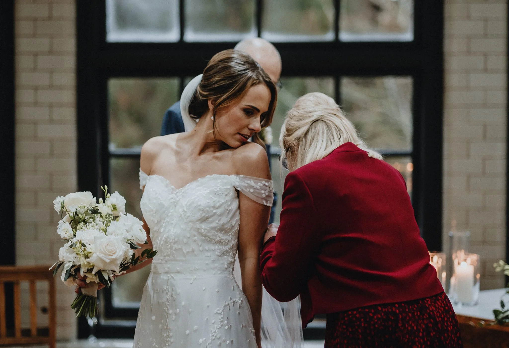Bride in a white wedding dress holding a bouquet of white flowers, leaning toward an older woman in a red blazer, in a warmly lit indoor setting.