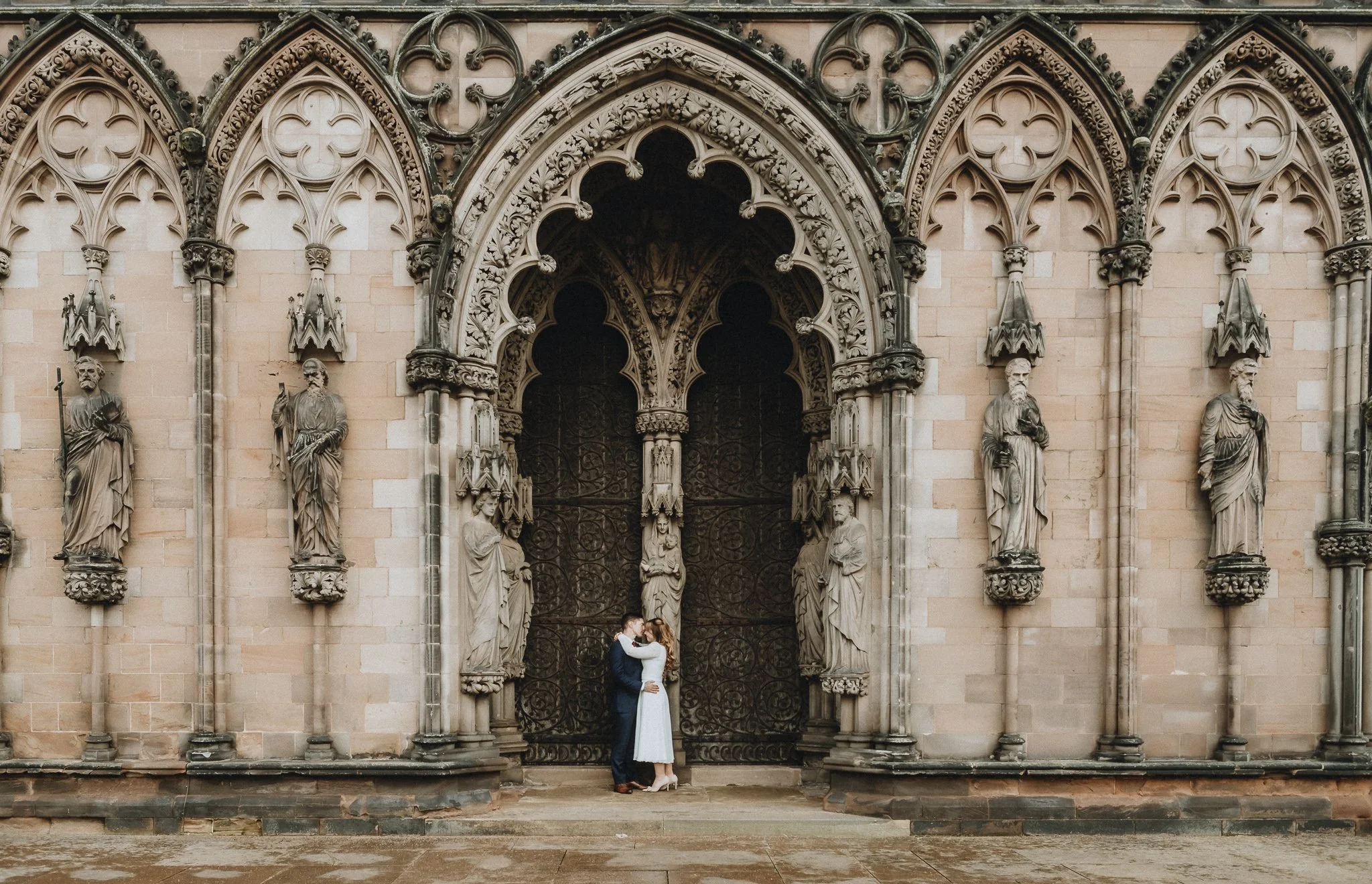 A couple dressed in wedding attire hugging in front of ornate gothic cathedral entrance with intricate stone carvings and statues.