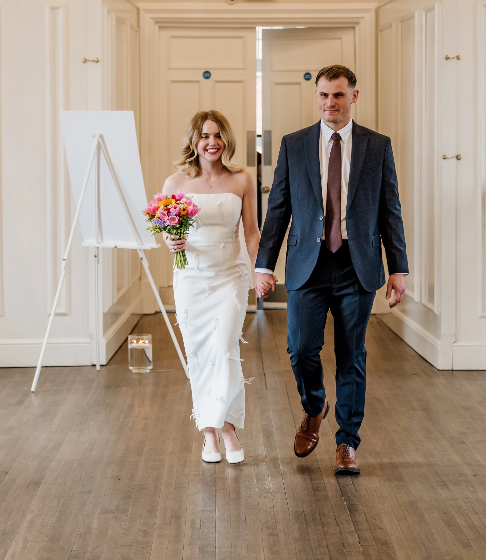 A bride and groom walking hand in hand indoors, smiling, with the bride holding a bouquet of pink and yellow flowers. The bride is wearing a white strapless dress and white heels, and the groom is wearing a dark suit with a white shirt and a brown ti