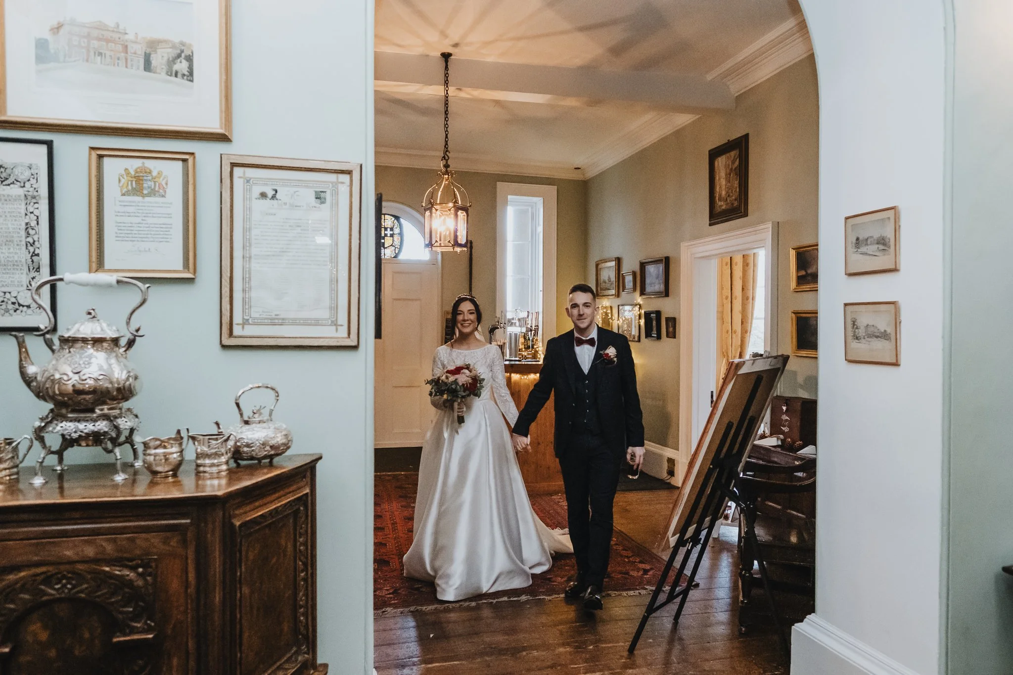 A bride and groom walking hand in hand inside a decorated house, smiling. The bride wears a white wedding gown and holds a bouquet, the groom wears a black suit with a bow tie. The interior features framed artwork on the walls, a wooden sideboard wit