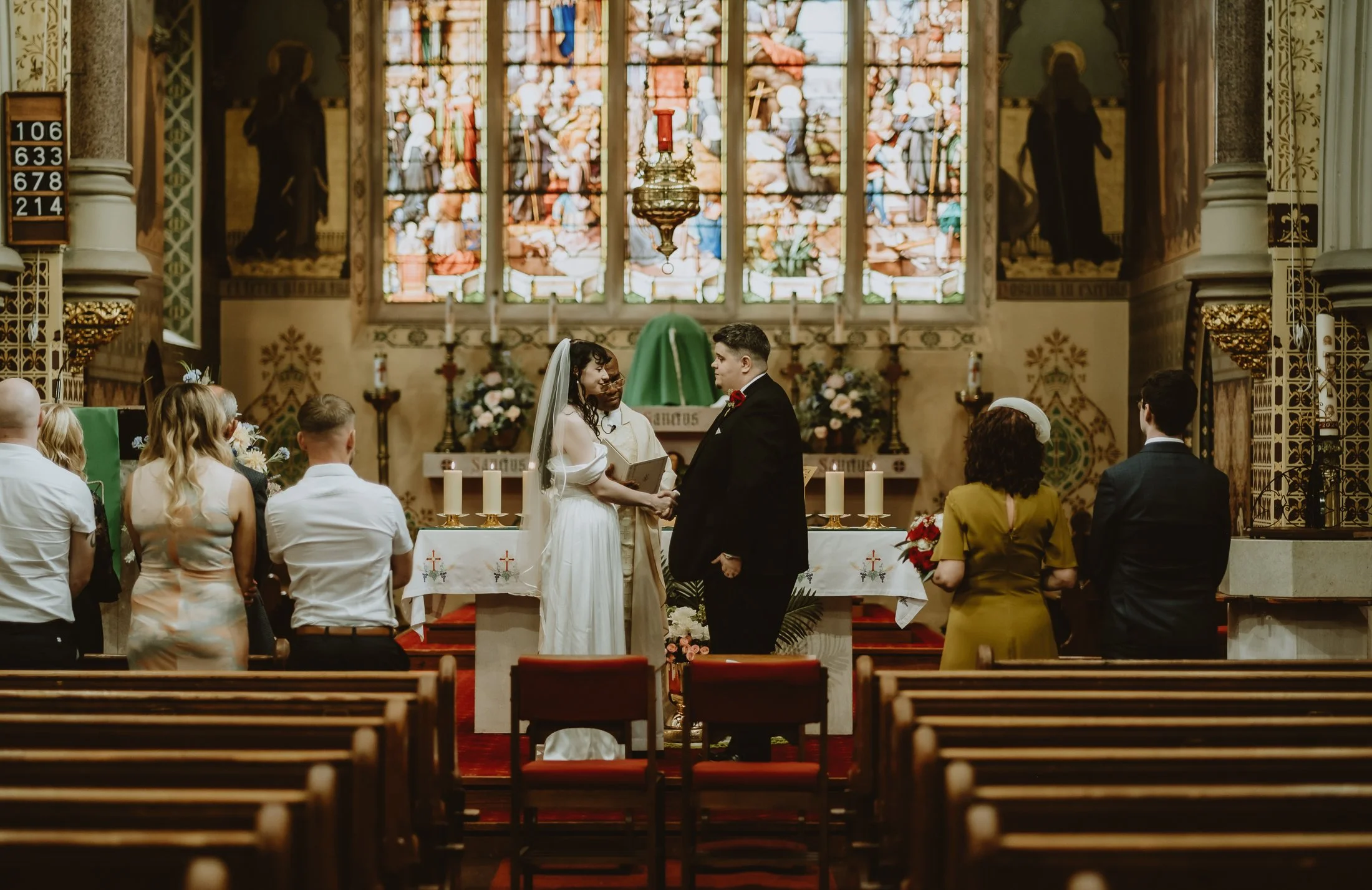 A wedding ceremony taking place in a church with a bride and groom holding hands at the altar, surrounded by guests, with stained glass windows and religious icons in the background.