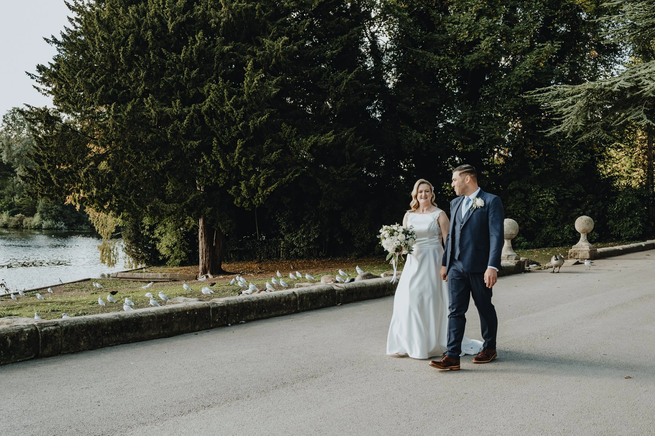 A bride and groom walking outdoors near a river, with tall green trees and birds on the ground.