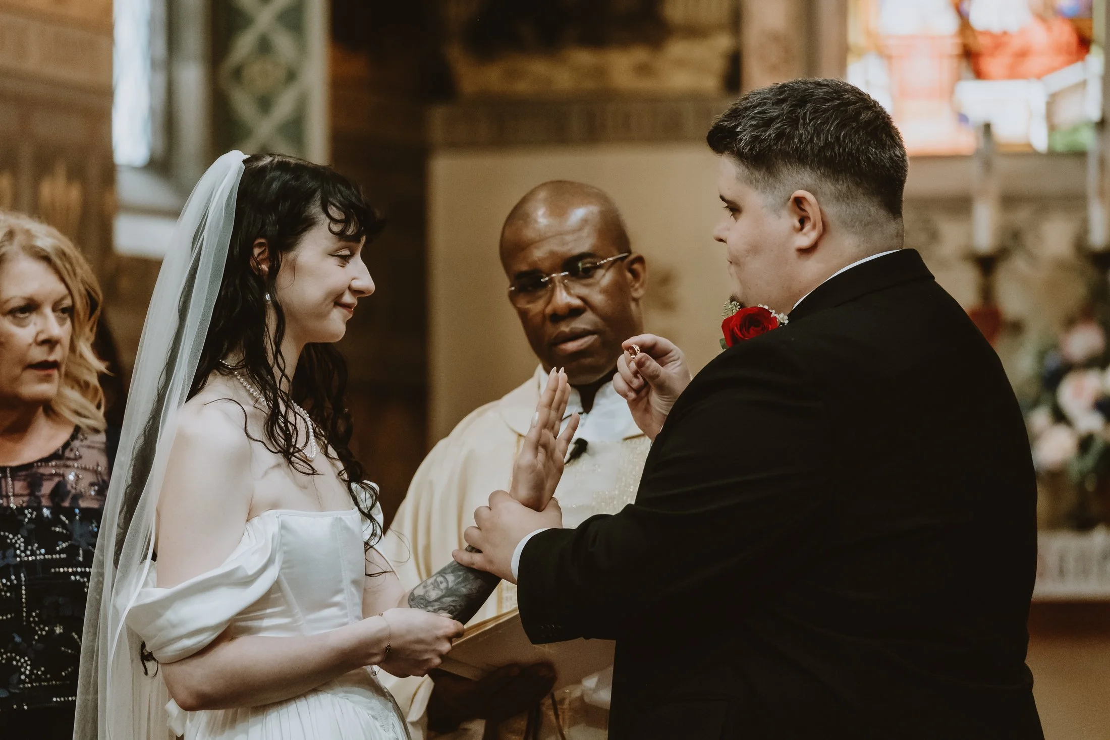A bride and groom exchange wedding vows and rings during a wedding ceremony in a church, with an officiant and a guest present.