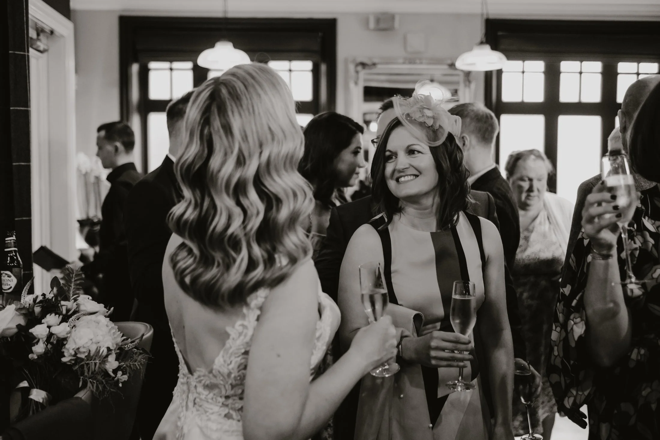 A group of people attending a social gathering, some holding glasses of champagne, in a warmly lit indoor space with large windows.