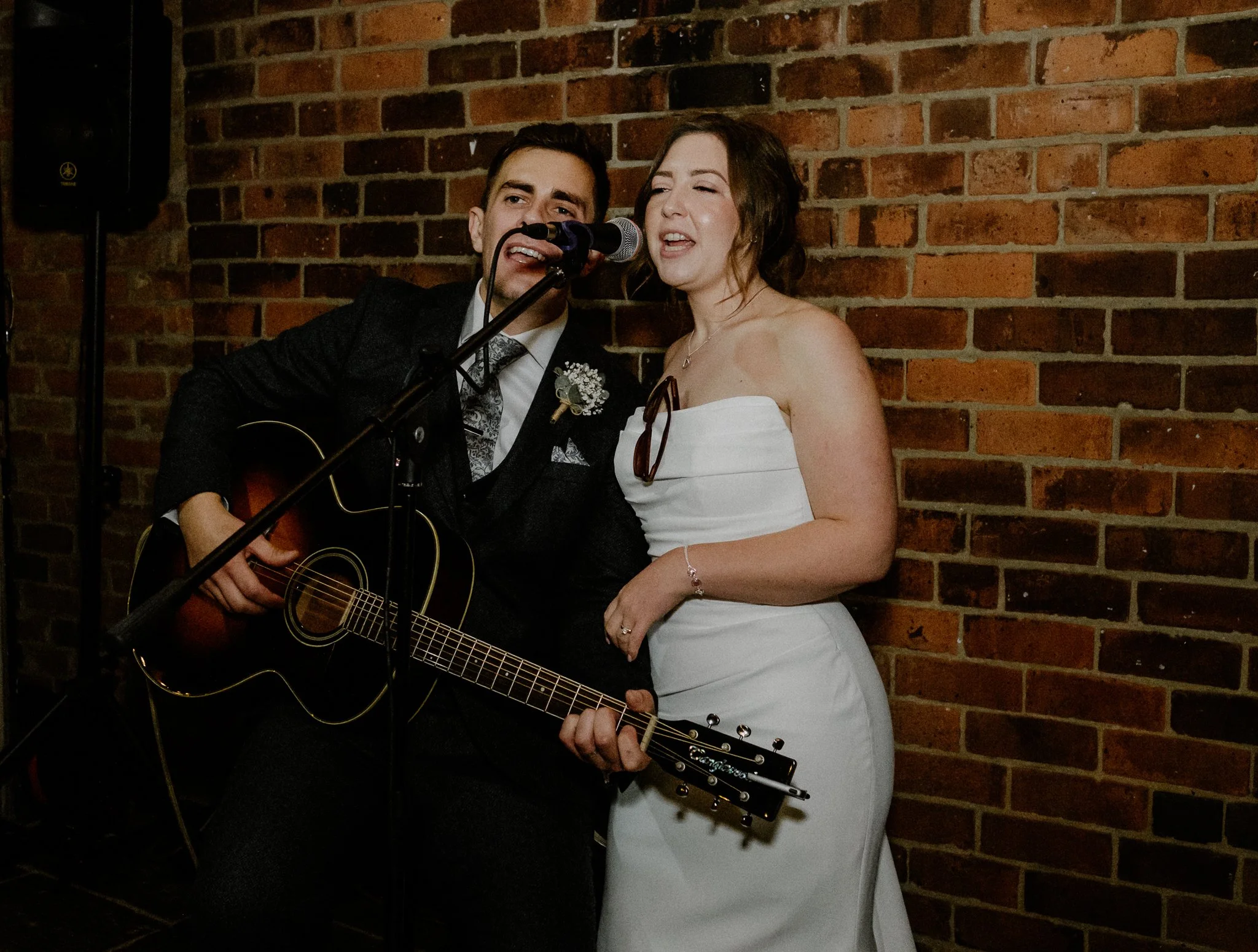 A man in a tuxedo playing guitar and singing into a microphone, with a woman in a white strapless dress singing next to him, against a brick wall.