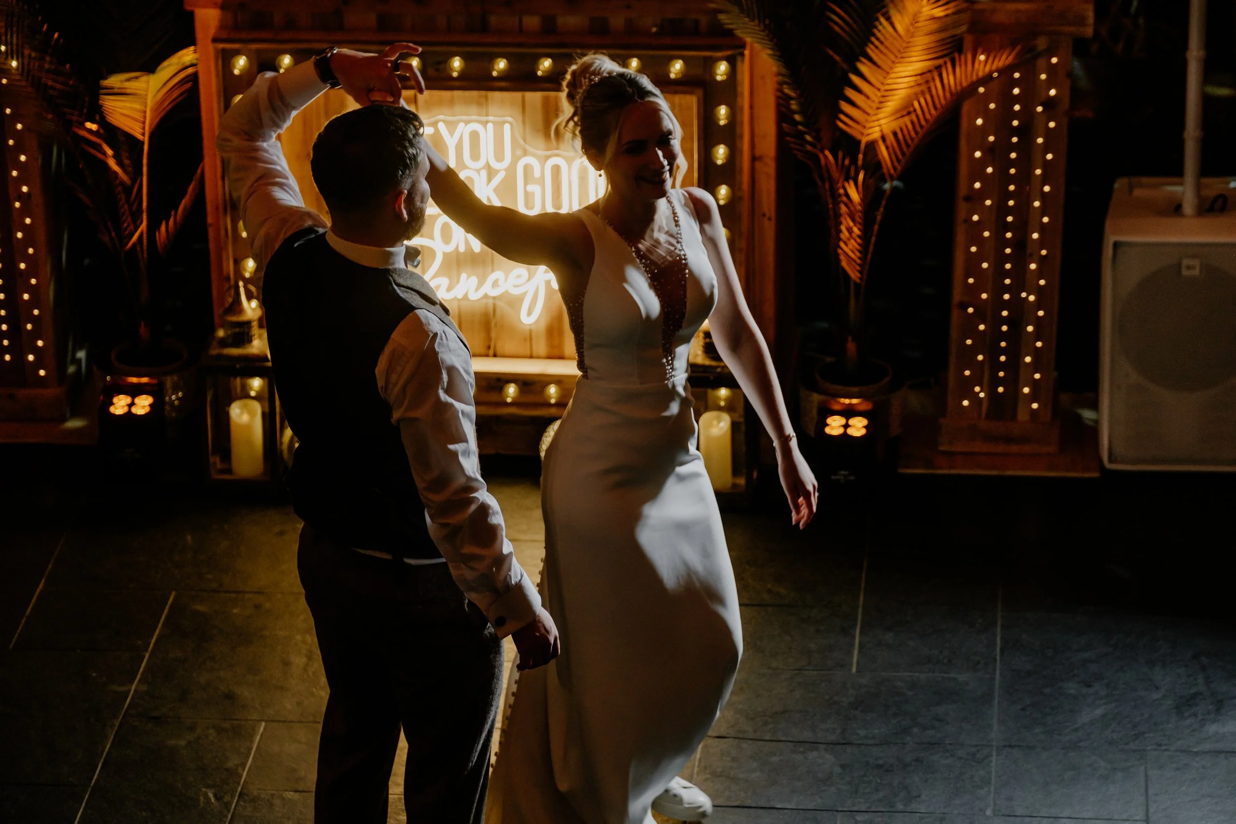 A bride and groom dancing at their wedding reception, with the bride wearing a white dress and the groom in a dark vest, in a decorated venue with a neon sign and candles in the background.