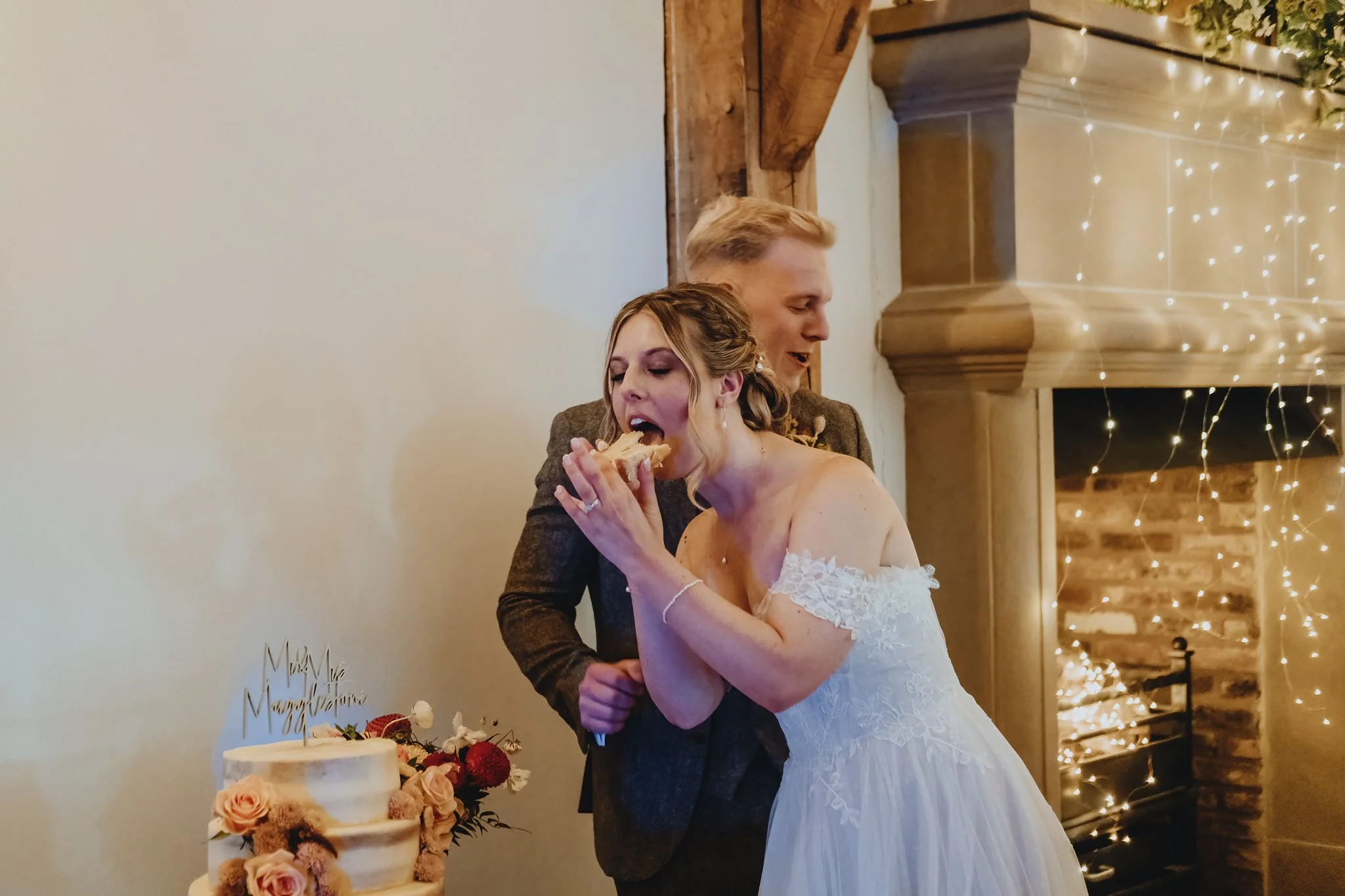 Bride and groom at wedding cake cutting, bride in white lace dress, groom in suit, standing near decorated wedding cake with flowers, fireplace with fairy lights in background.