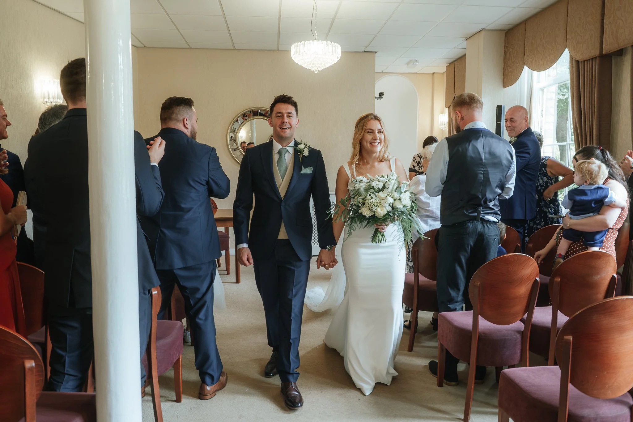 A newlywed couple walking hand in hand down the aisle of a wedding ceremony, surrounded by guests. The bride is holding a bouquet and smiling, while the groom is dressed in a suit and also smiling. Guests are clapping and celebrating in a decorated i