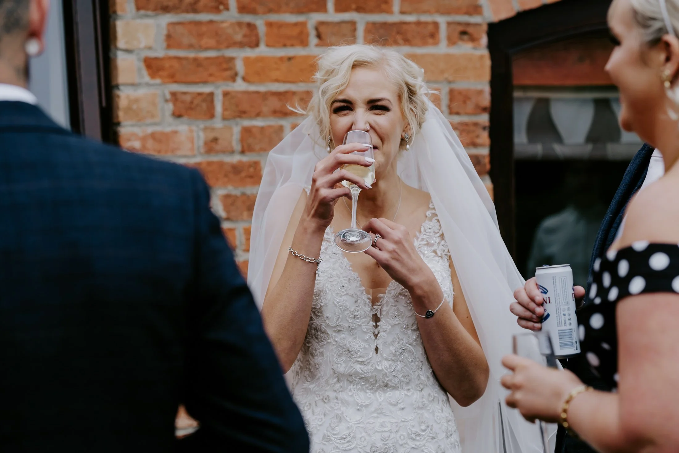 A bride wearing a white wedding dress and veil is drinking from a champagne flute. She is surrounded by guests, one holding a soda can. The background features a brick wall.