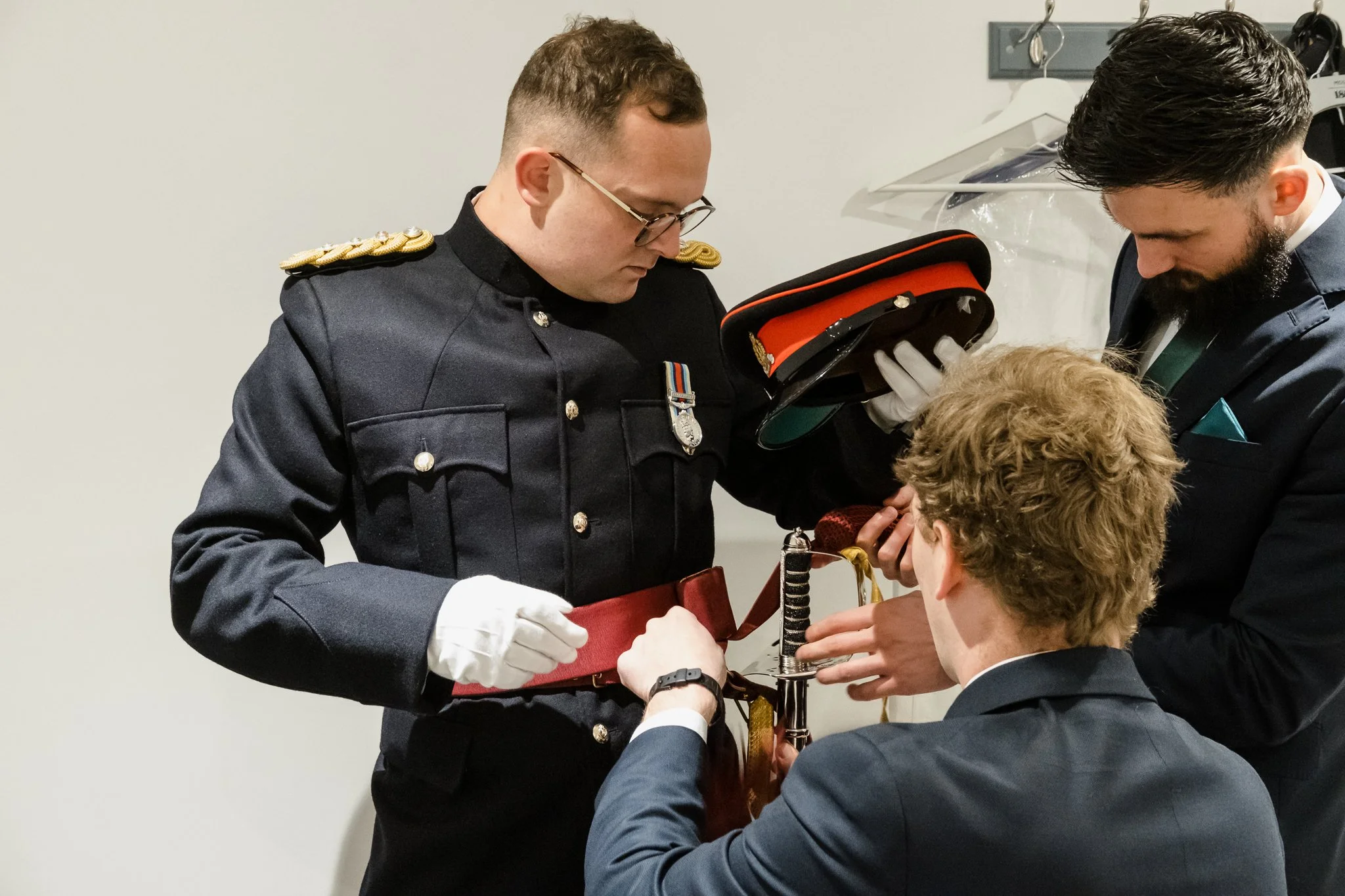 Three men, two in formal suits and one in a military uniform, are engaged in a ceremonial activity involving a sword. The man in uniform is holding a hat, while the other two men are assisting with the sword, which appears to be part of an official o