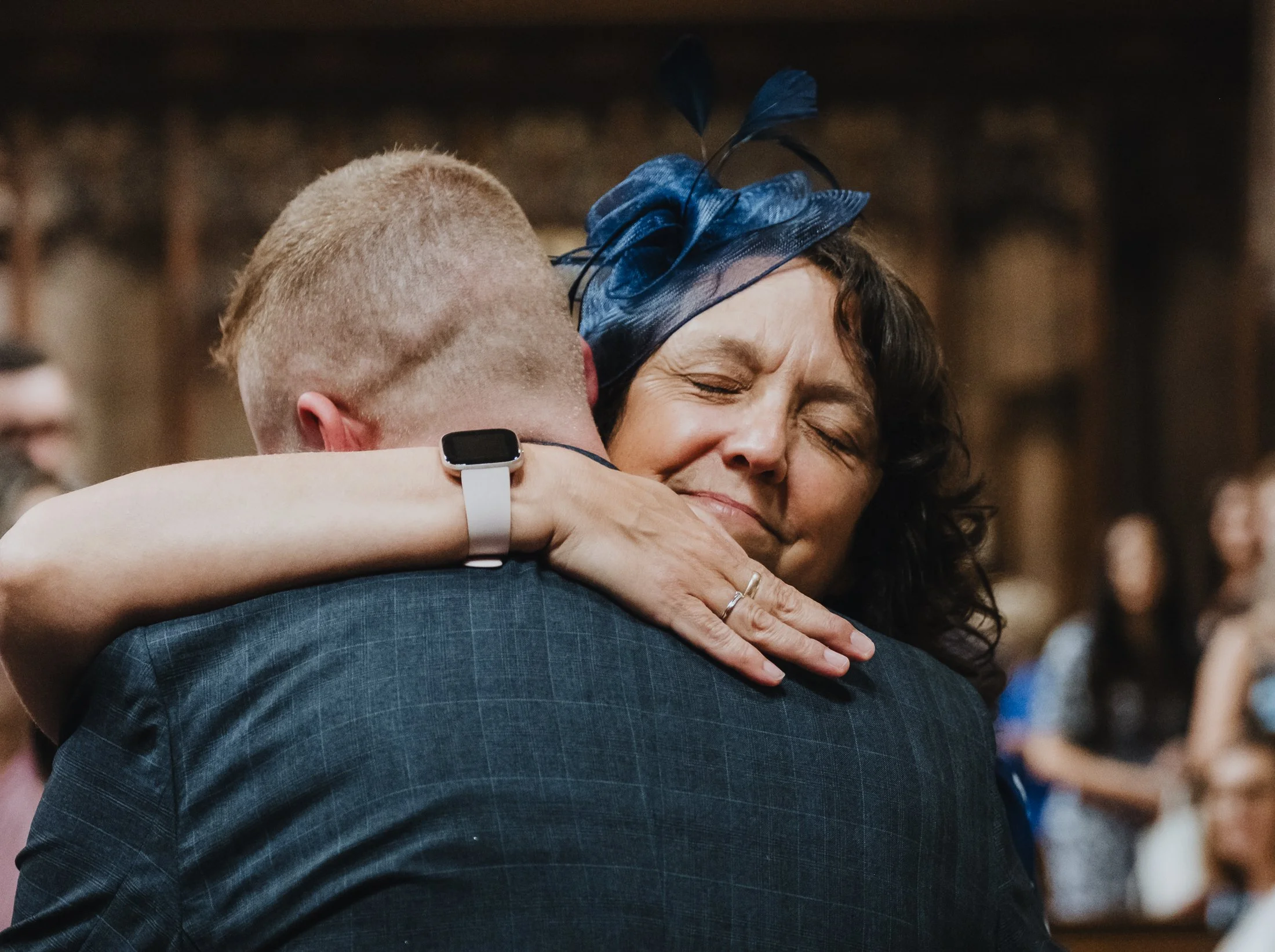 A woman with curly dark hair and a blue fascinator hat hugging a man in a dark blue suit at what appears to be a wedding or similar celebration.