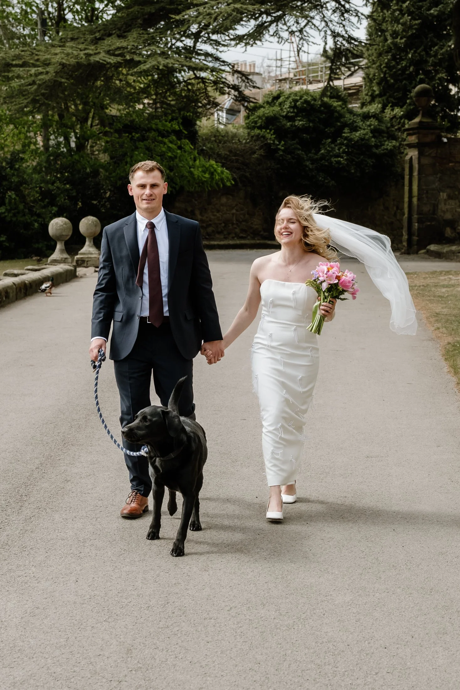 A bride and groom walking hand in hand with a black dog on a leash in a park. The bride is wearing a white strapless wedding dress and holding a bouquet of pink flowers, with a veil flowing in the wind. The groom is wearing a navy suit, white shirt, 