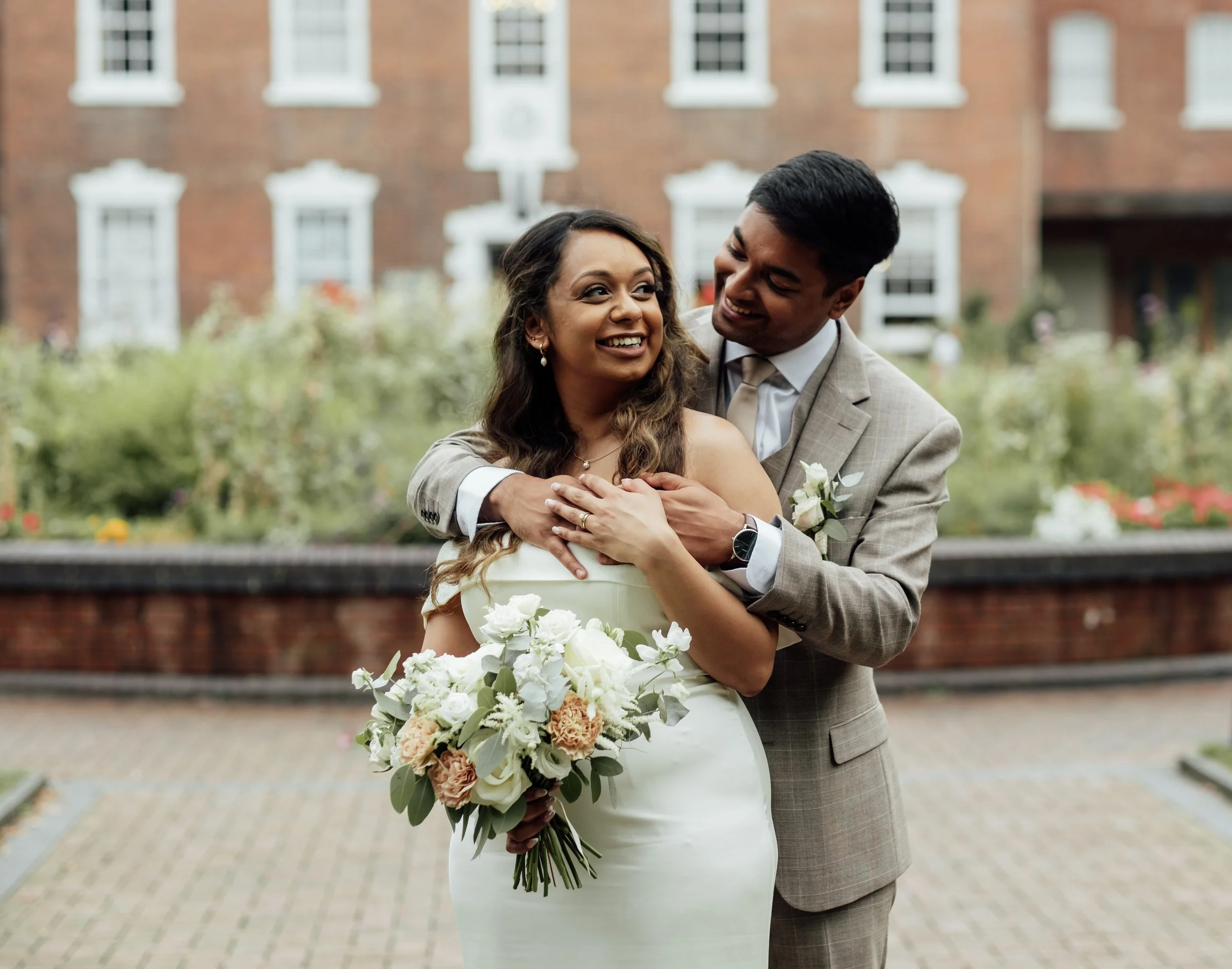 A newlywed couple is hugging and smiling outdoors in front of a brick building. The woman is holding a bouquet of white and blush roses, and the man is wearing a beige checked suit with a boutonniere. They look happy and affectionate.