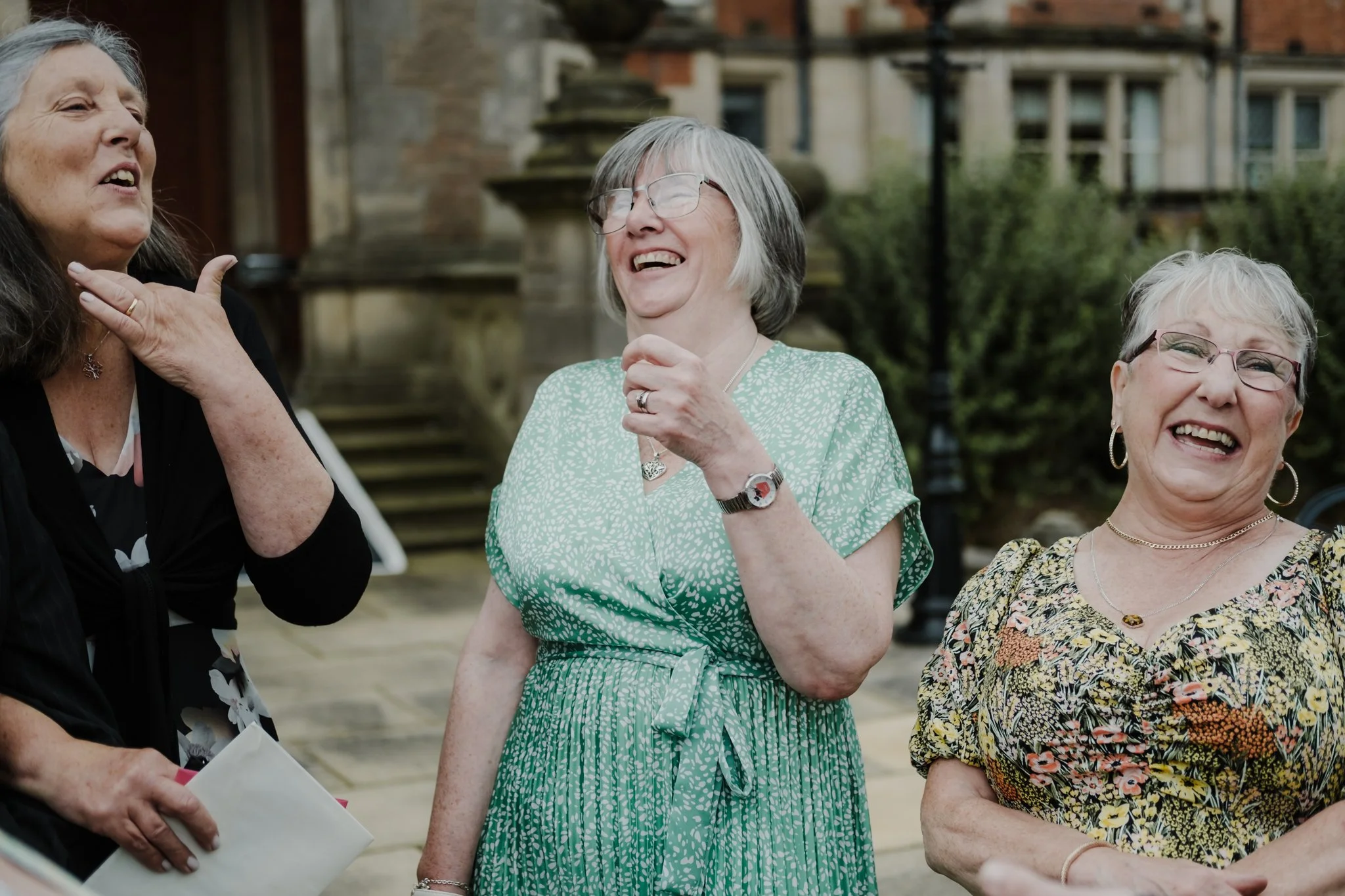 Three elderly women laughing and talking outdoors in front of historic buildings, one wearing glasses and a green dress, another with short gray hair and a floral dress, the third partially visible wearing a black top.