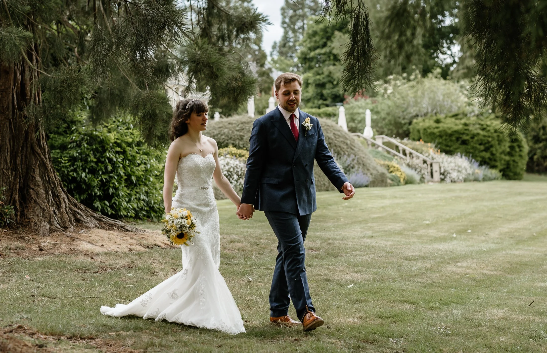 A bride and groom walking hand in hand through a lush garden, with trees and shrubs in the background, celebrating their wedding day.