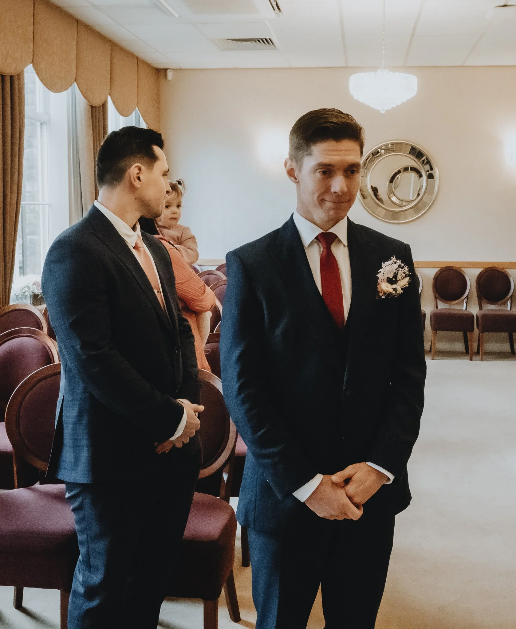 Three men and a little girl, all dressed in formal attire, in a decorated room with chairs, a chandelier, and a round wall mirror.