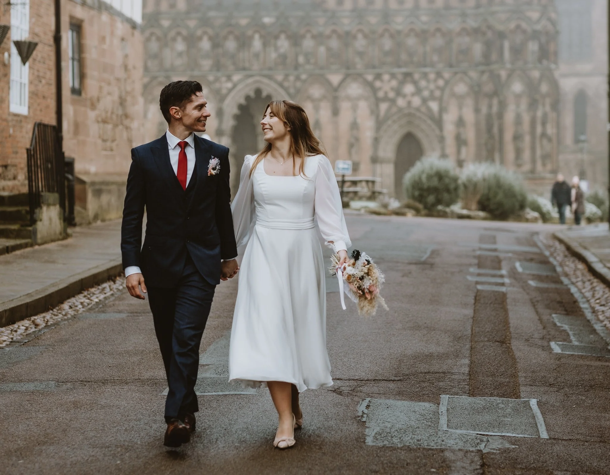 A bride and groom walking hand in hand on a city street, smiling at each other, with a historic cathedral in the background; the bride holds a bouquet of flowers.