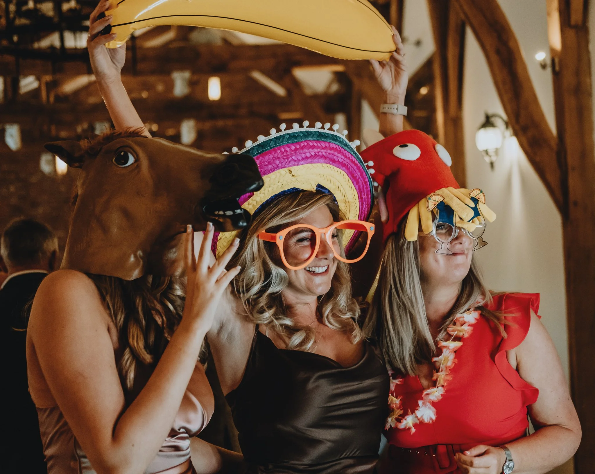 Two women at a party wearing fun hats and oversized glasses, one holding a horse mask and the other with a red hat with cartoon eyes and a yellow decoration, smiling and posing.