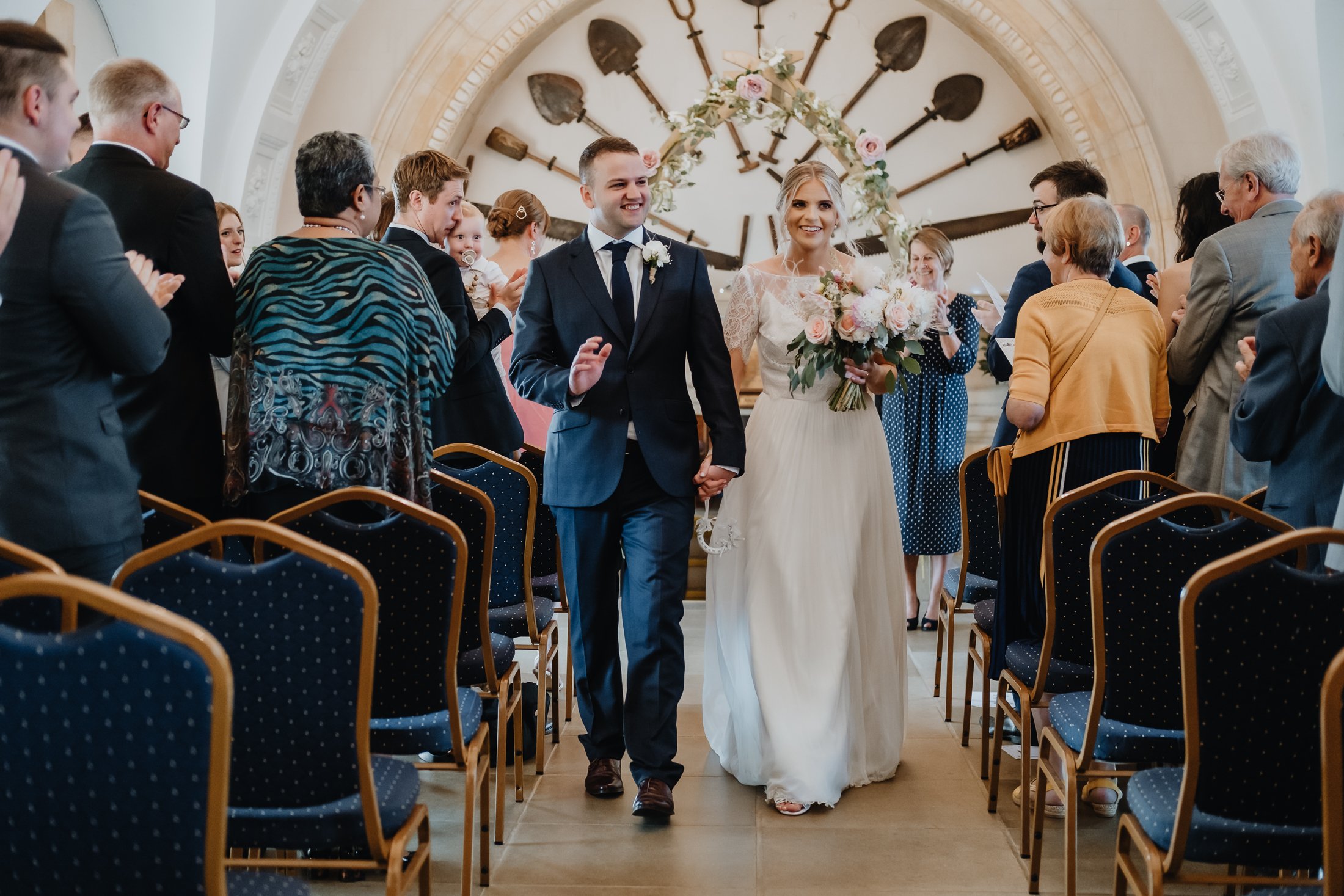 A bride and groom walk down the aisle at their wedding ceremony, smiling and holding hands, surrounded by seated guests clapping and celebrating indoors.