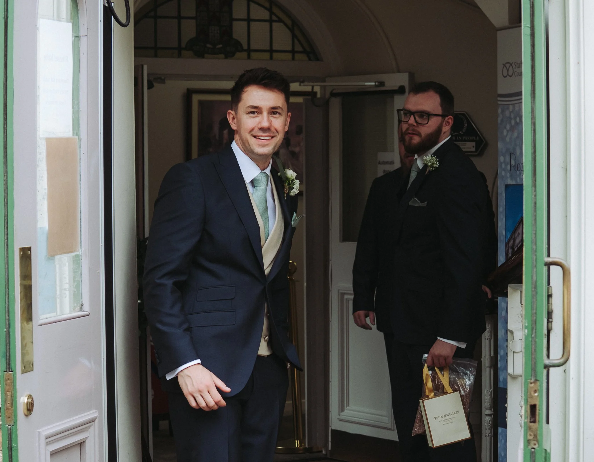 Two men in suits standing at the doorway of a building, one smiling at the camera and the other holding a gift bag, both wearing boutonnieres, during a formal event.