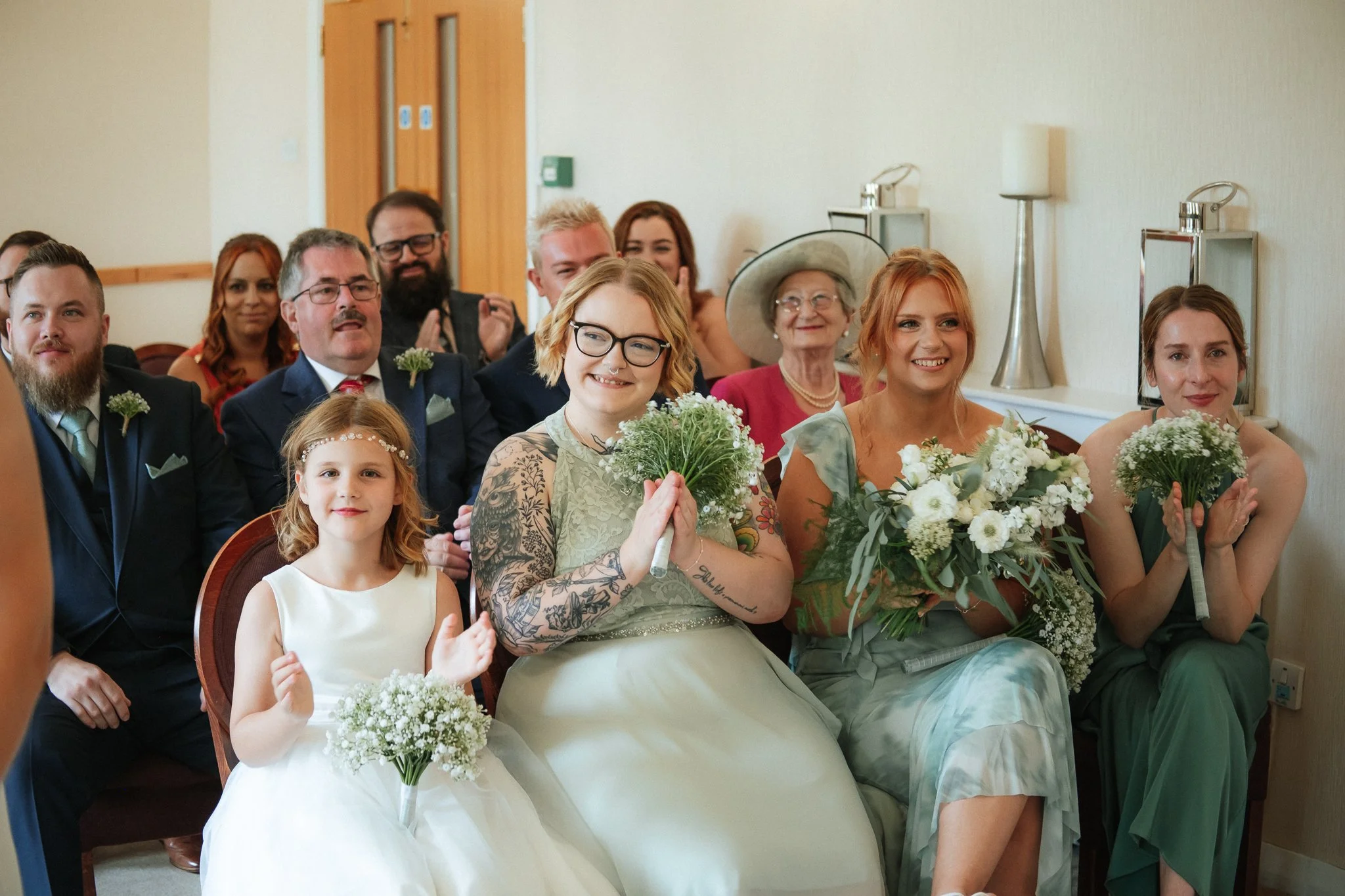 A group of people sitting in a wedding ceremony, including women with bouquets, a young girl in a white dress, and several men in suits.
