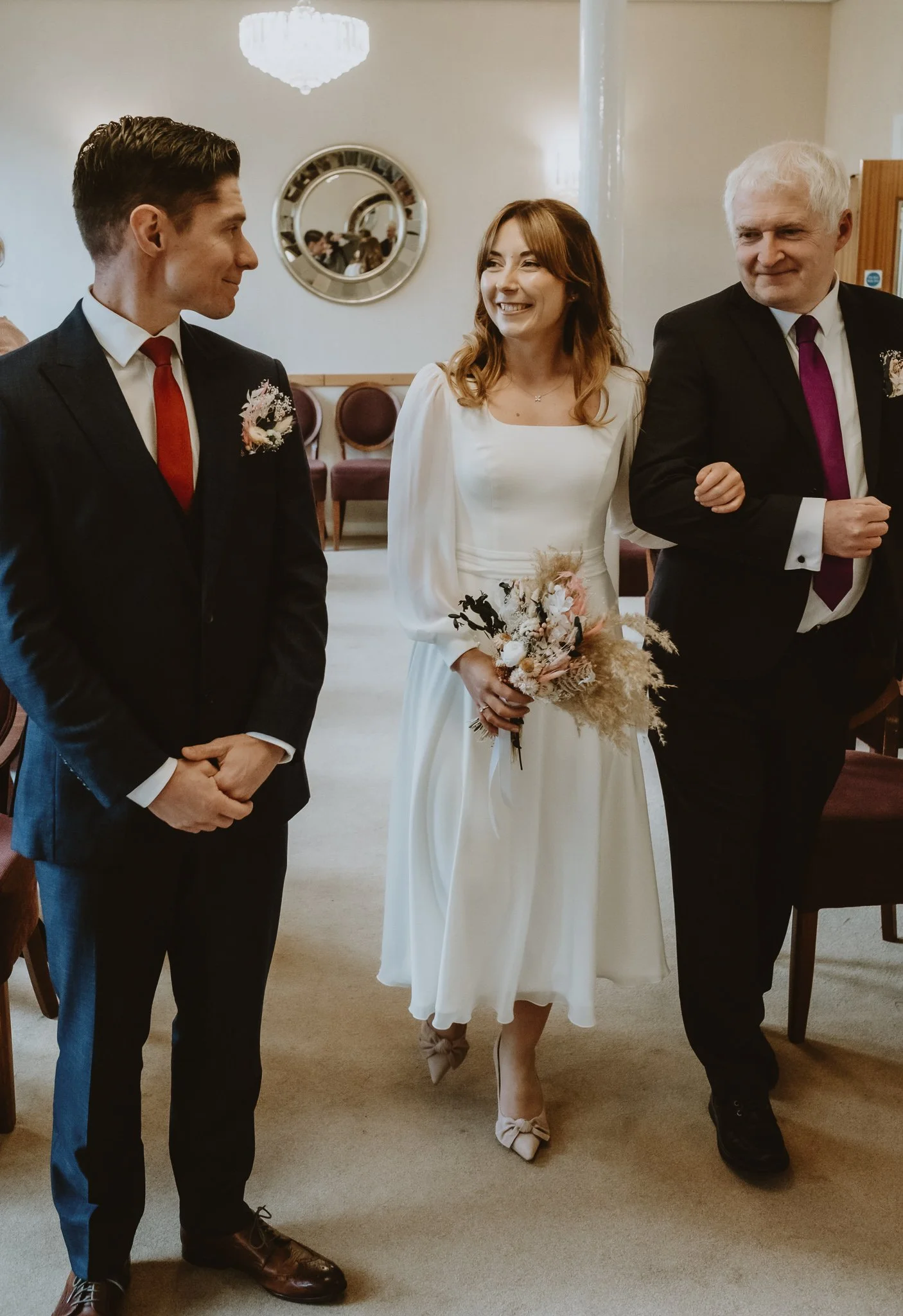 A bride in a white dress holding a bouquet, standing between two men in suits, in a room with a mirror and chandelier.