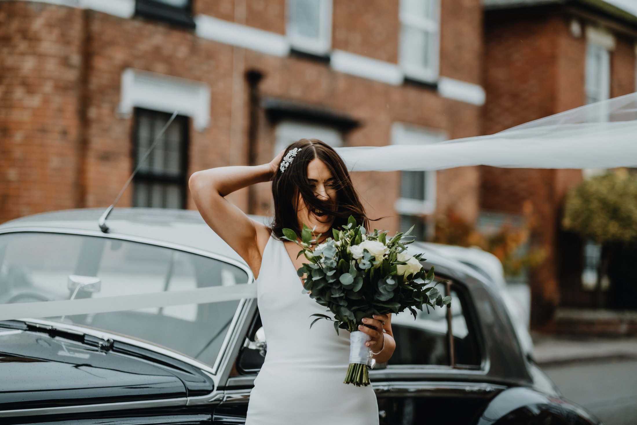 A smiling bride in a white wedding dress holding a bouquet of white flowers and greenery, next to a black vintage car with white ribbon decorations, in front of a brick building.
