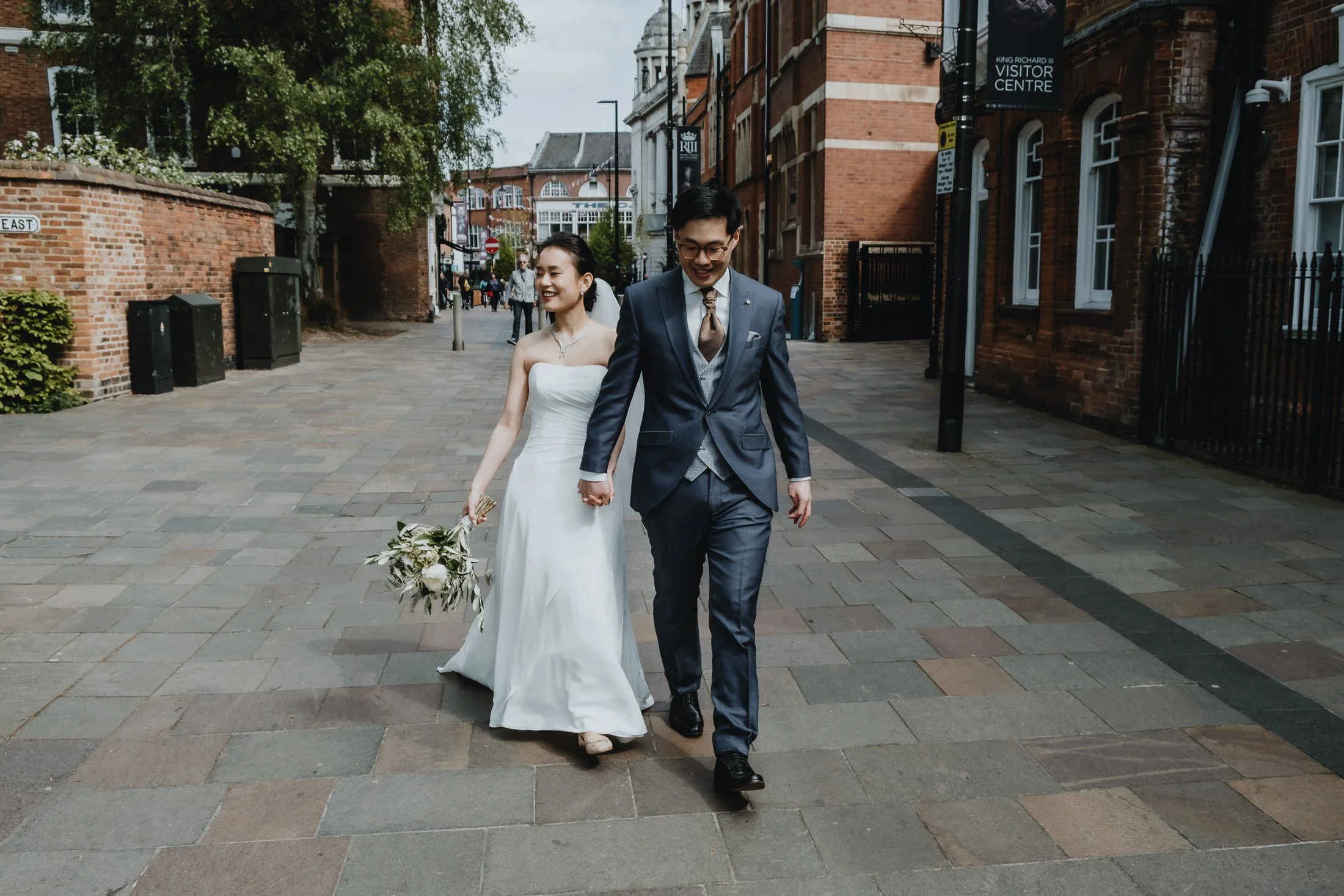 A newlywed couple walking hand in hand on a city street. The bride is wearing a white strapless wedding gown and holding a bouquet, while the groom is dressed in a dark suit and tie, smiling.