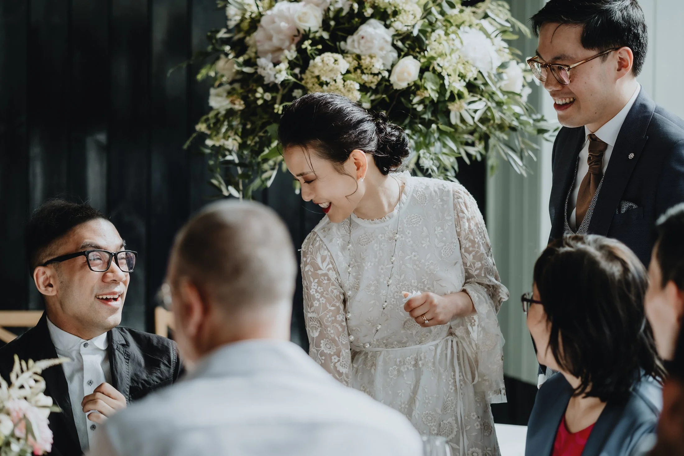 A group of friends at a celebration, laughing and smiling, with a woman in a white lace dress in the center and floral arrangements in the background.