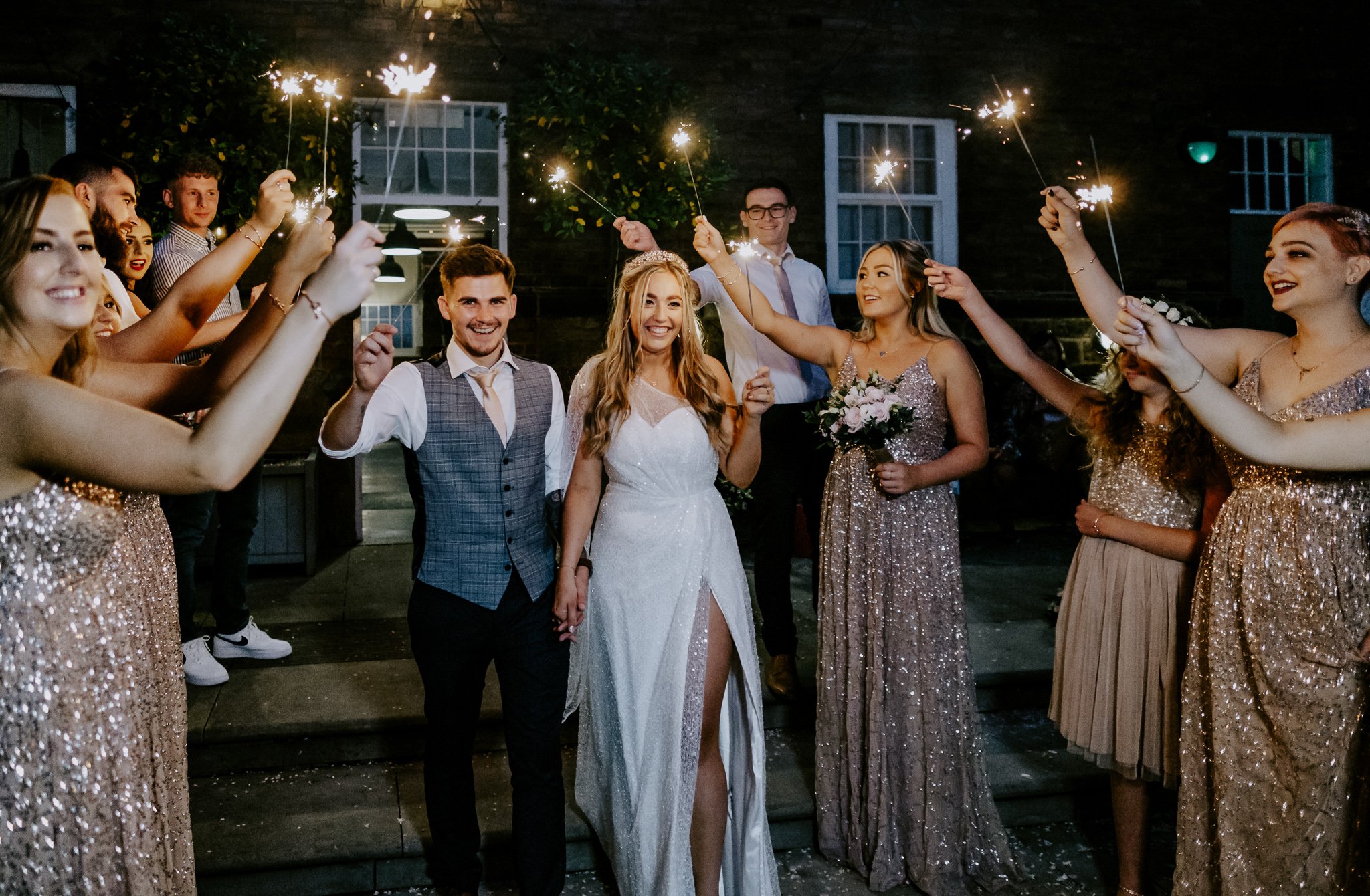 Group of people celebrating a wedding at night with sparklers, including a bride in a white dress, groom in a vest, and bridesmaids in sparkly dresses.